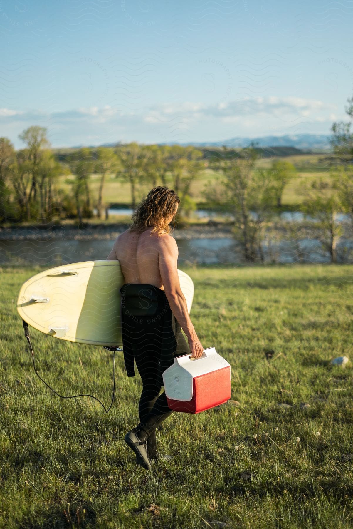 A man carries a surfboard and a cooler along a river in montana at dusk