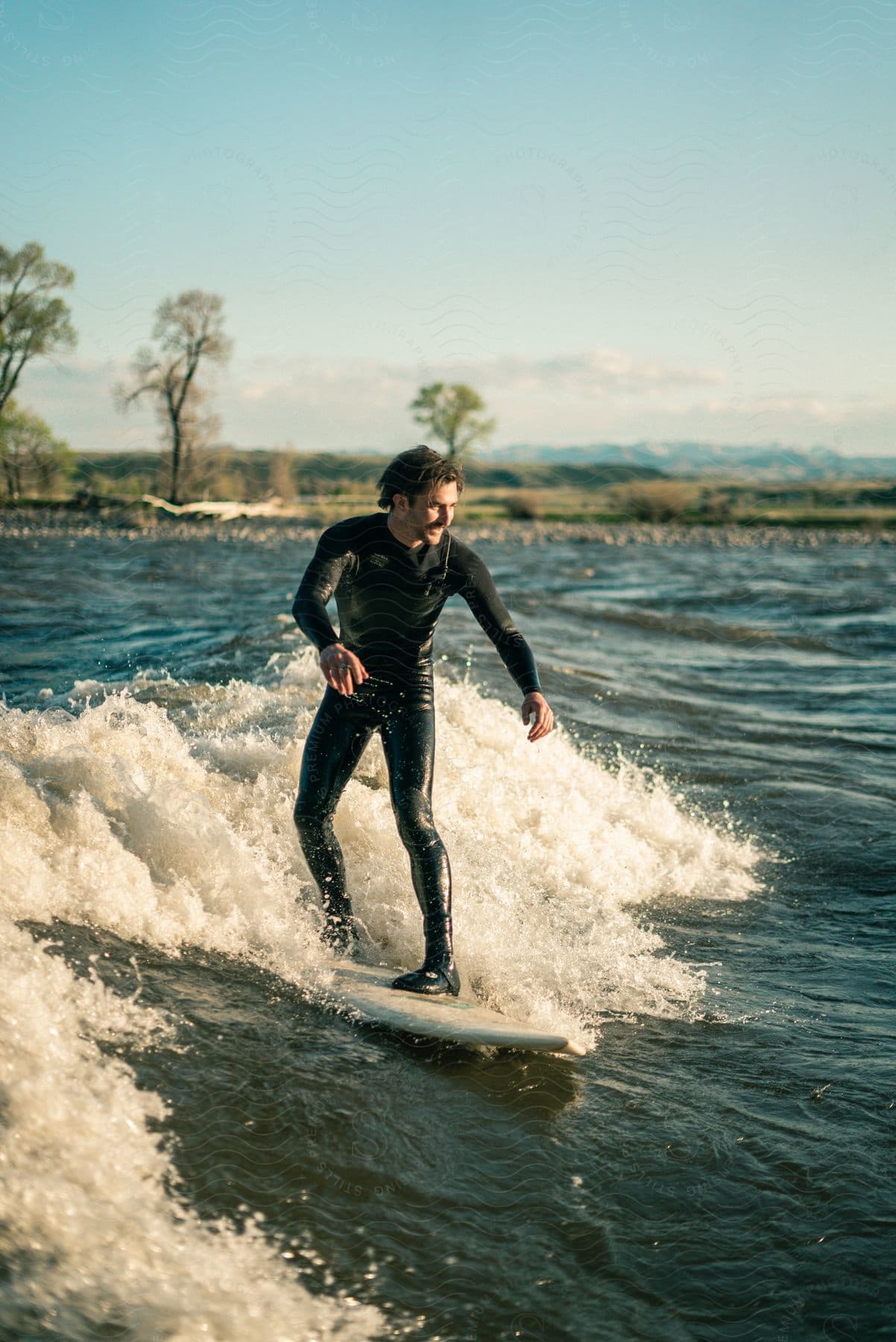 A man wearing a wetsuit is seen surfing on a standing river wave at dusk in montana