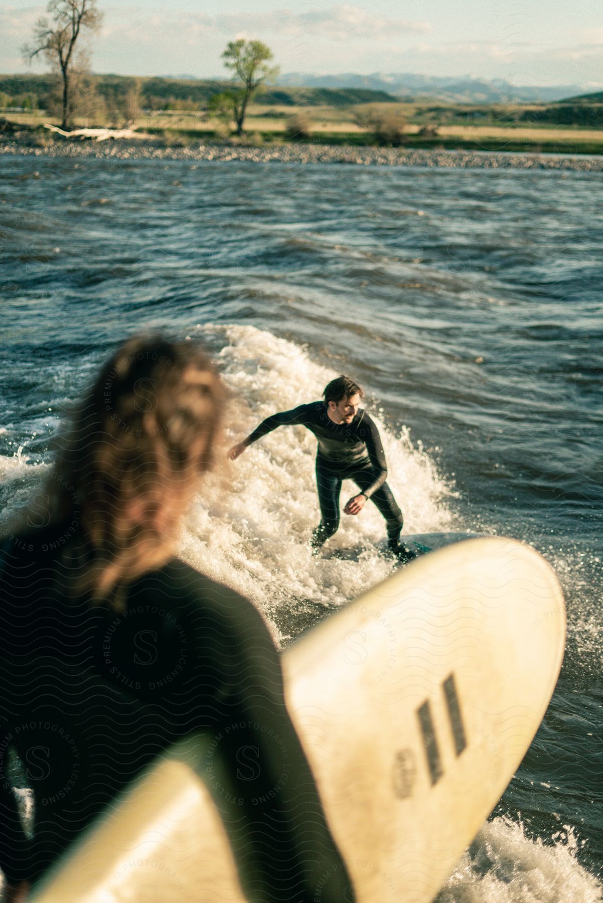 Person holding a surf board watching a man surf a wave with a landscape in the background