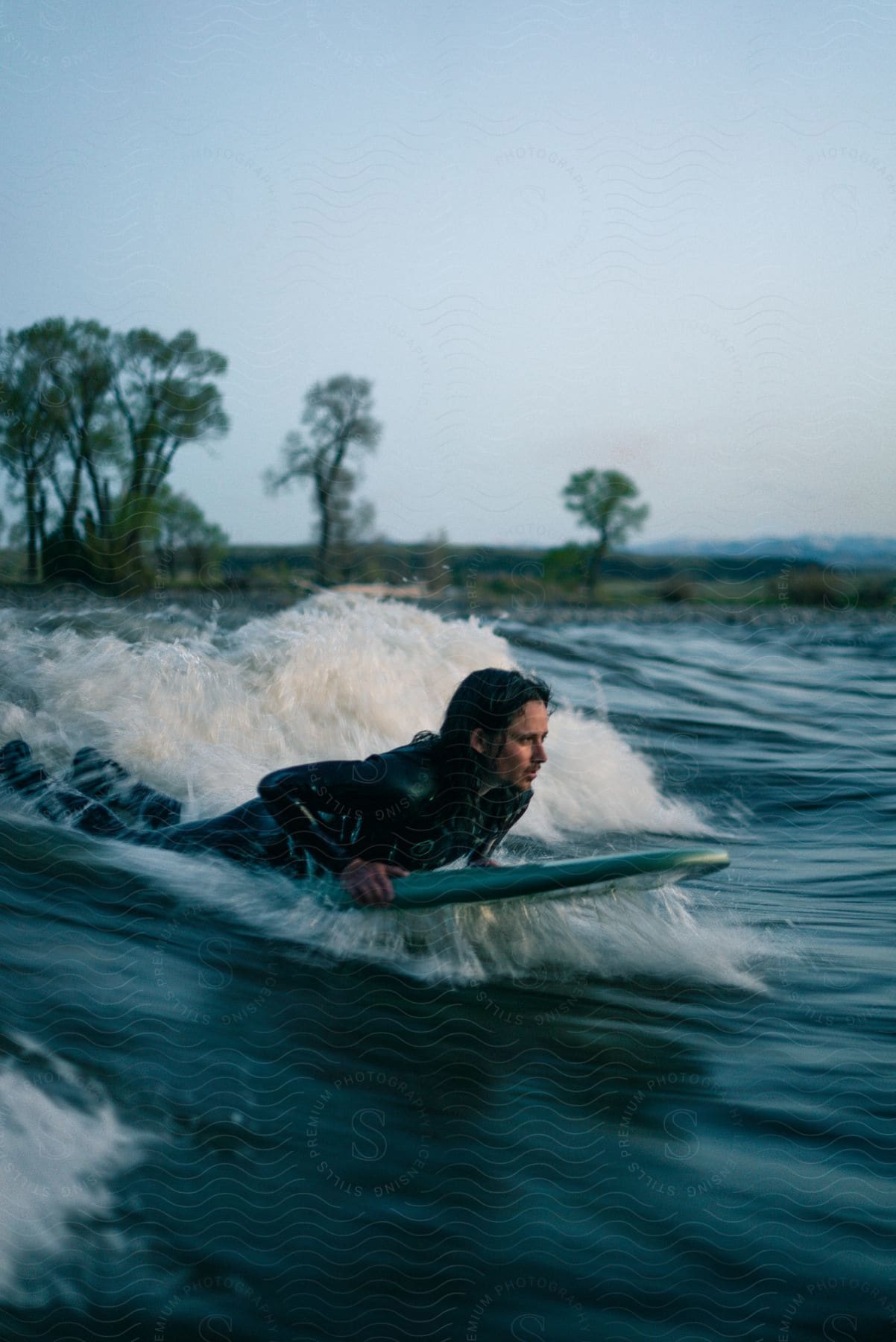 Man with long hair and wet suit paddles on surfboard in montana at dusk