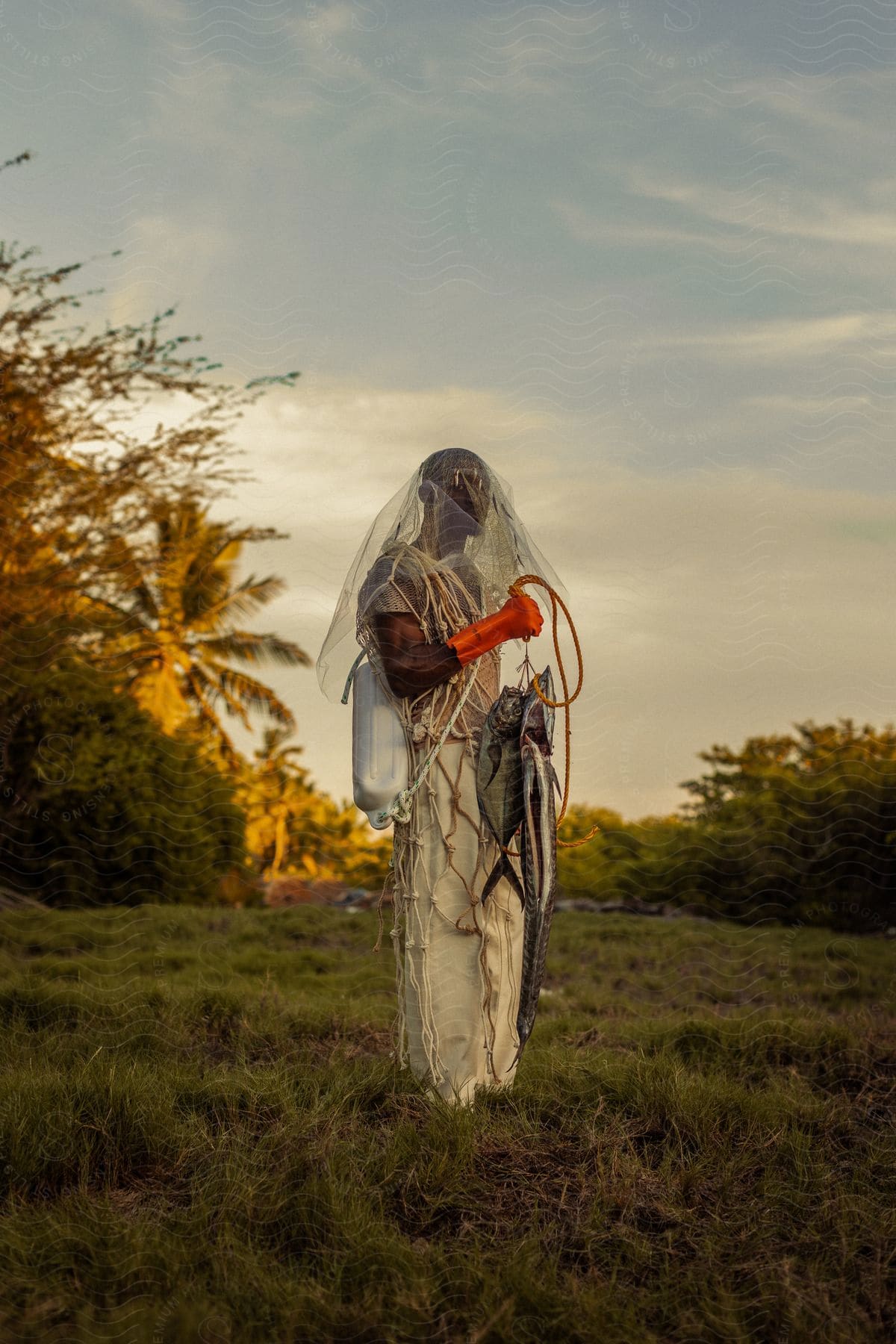 Man wearing white clothing and a bridal veil on his head, holding some fish in his hand in a natural outdoor setting in daylight.