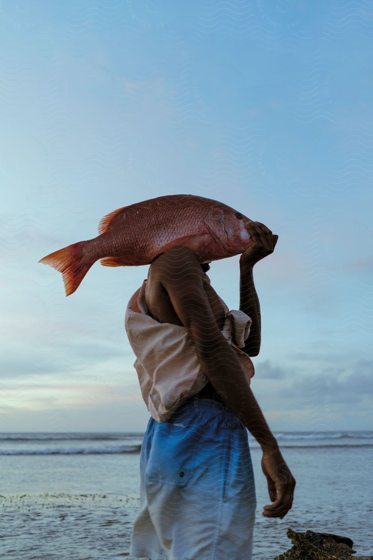 A man wearing a robe carries a large red fish over his shoulder on the beach