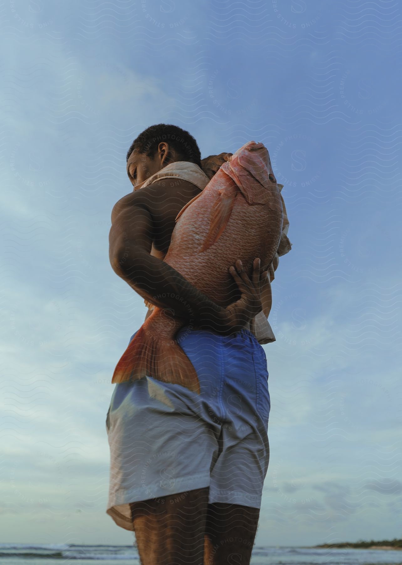 A man standing carrying a large red snapper on his back near the ocean