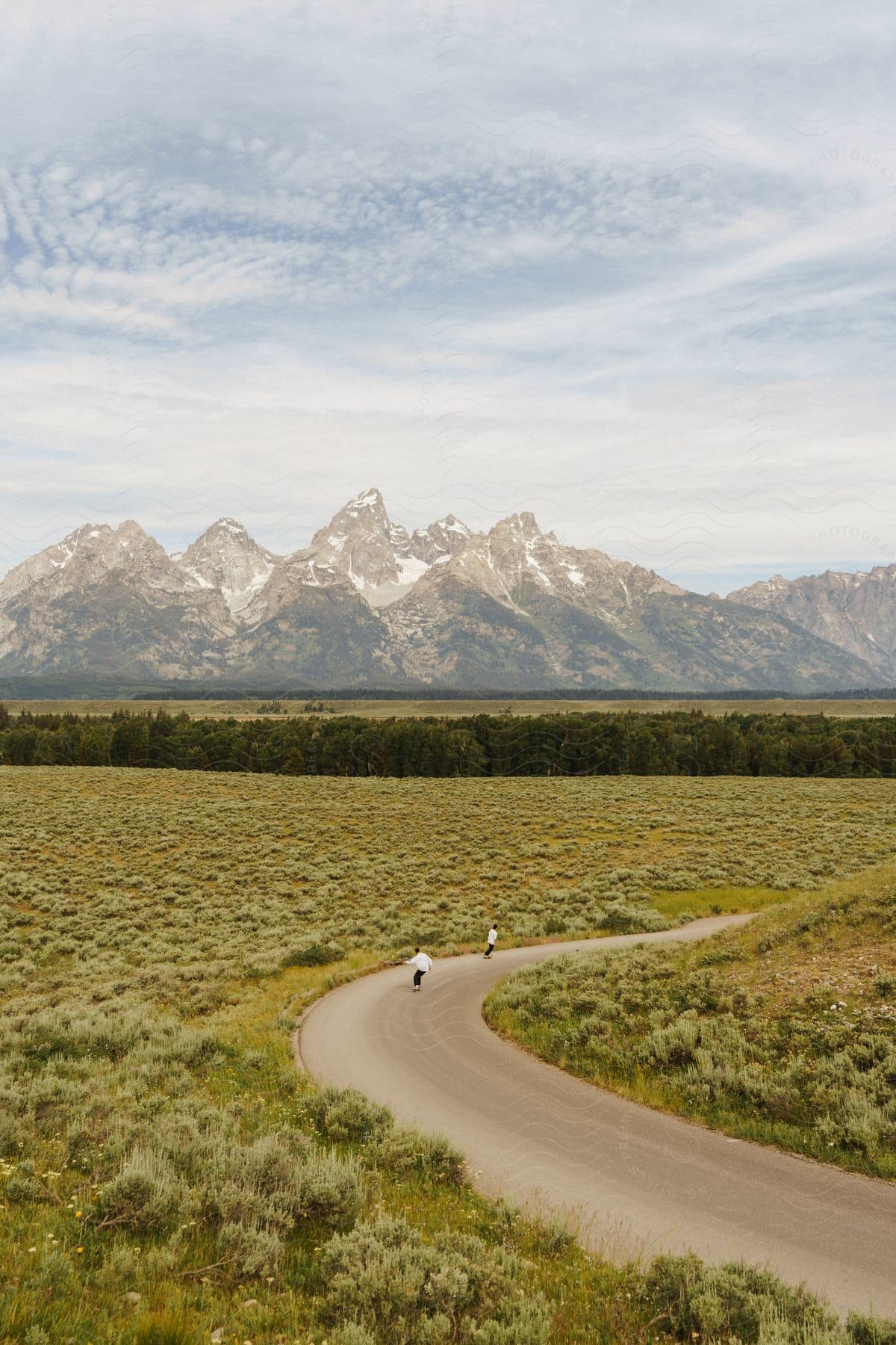 Skateboarders ride on a road near majestic mountains and a snow field surrounded by a dense forest