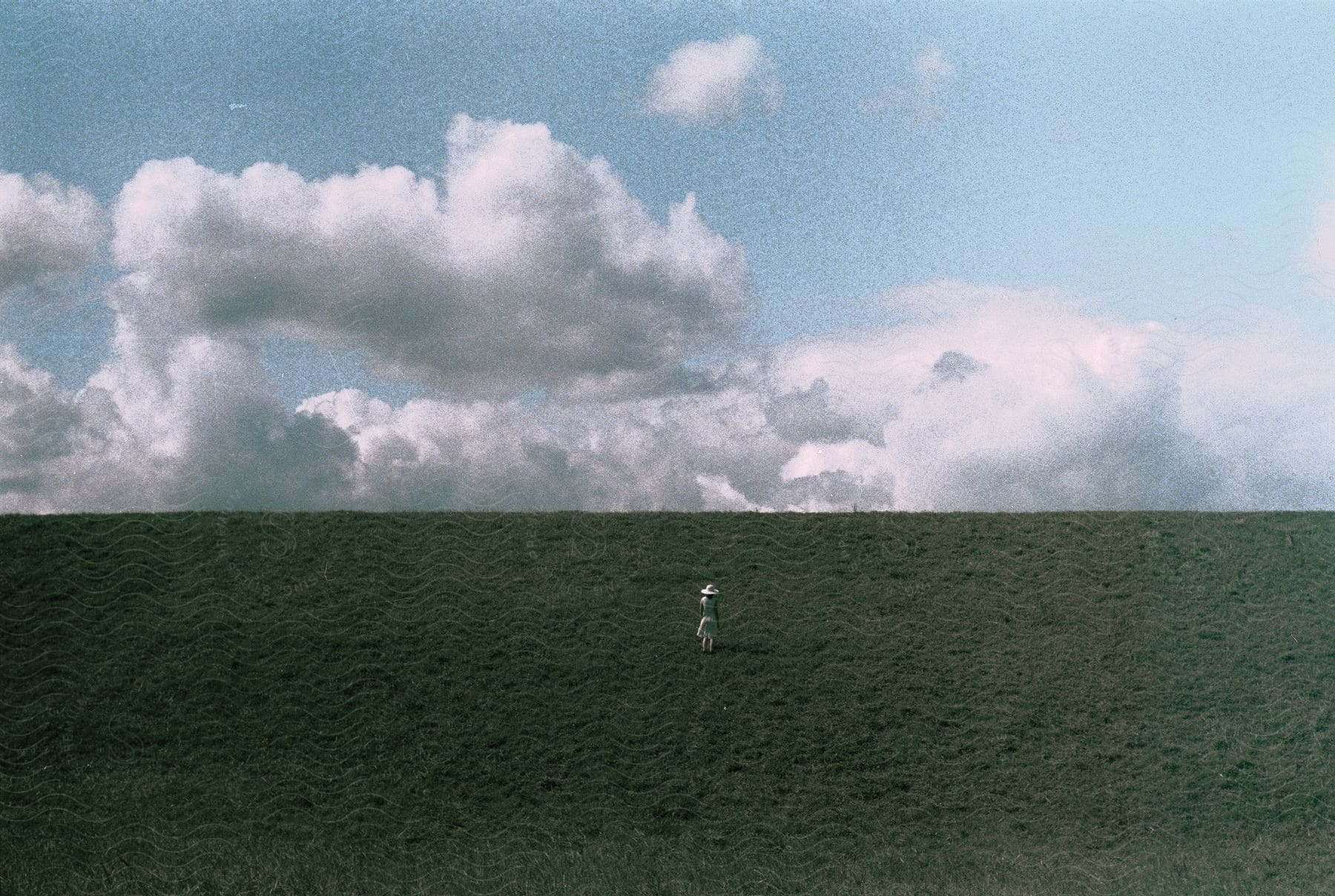 A distant woman wearing a sun hat and white dress walks uphill in a grassy landscape