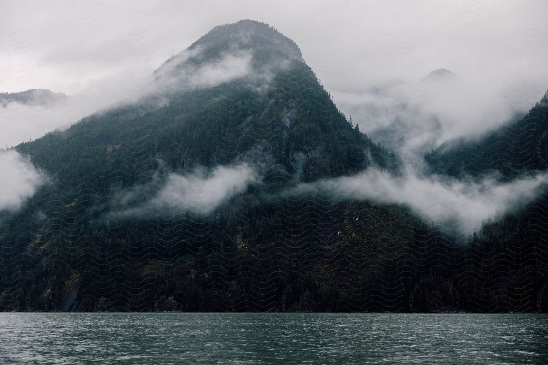 A treecovered mountain is surrounded by heavy clouds next to a calm sea