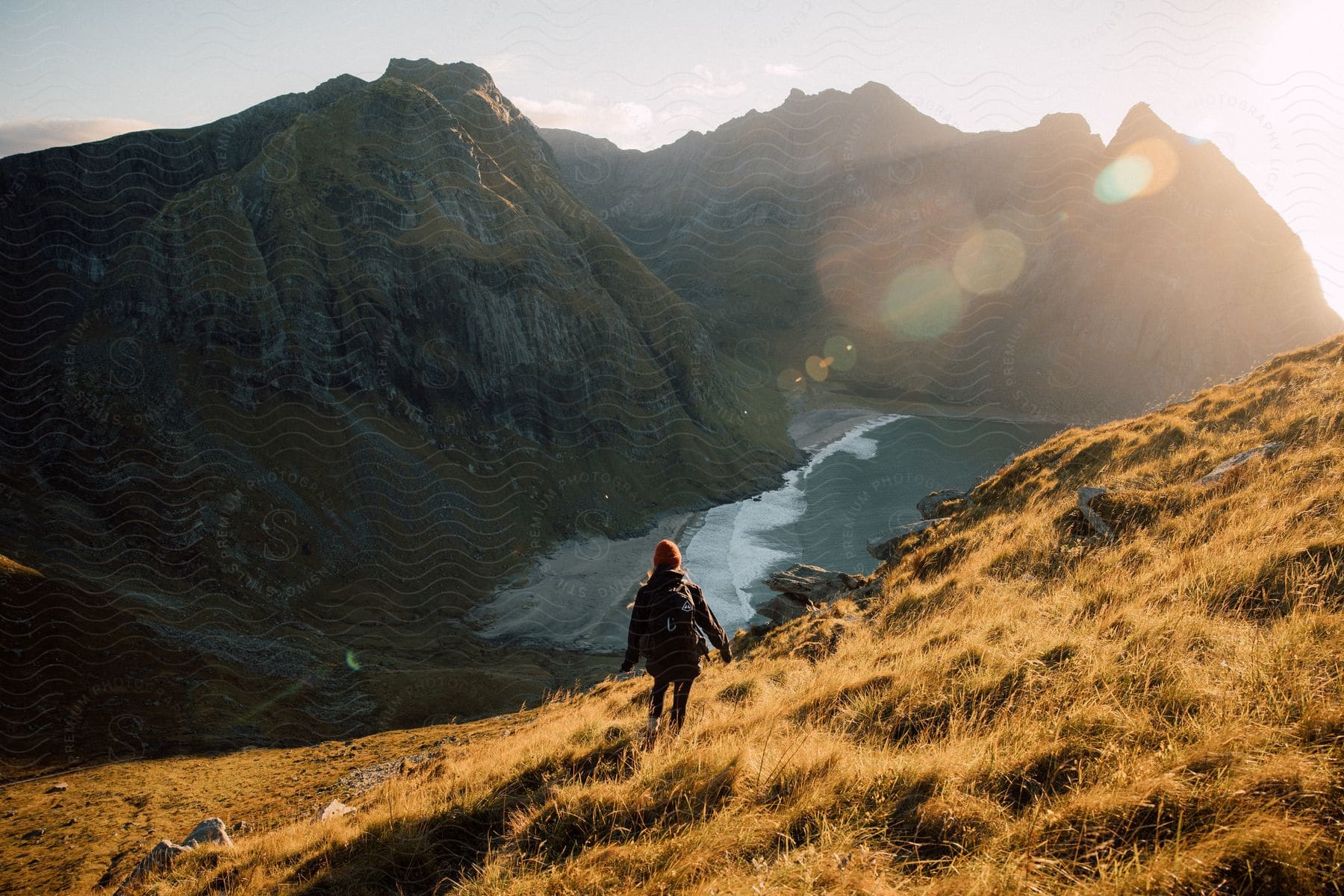 A woman with a backpack on a grassy hill overlooking water and rocky cliffs with the sun low in the sky