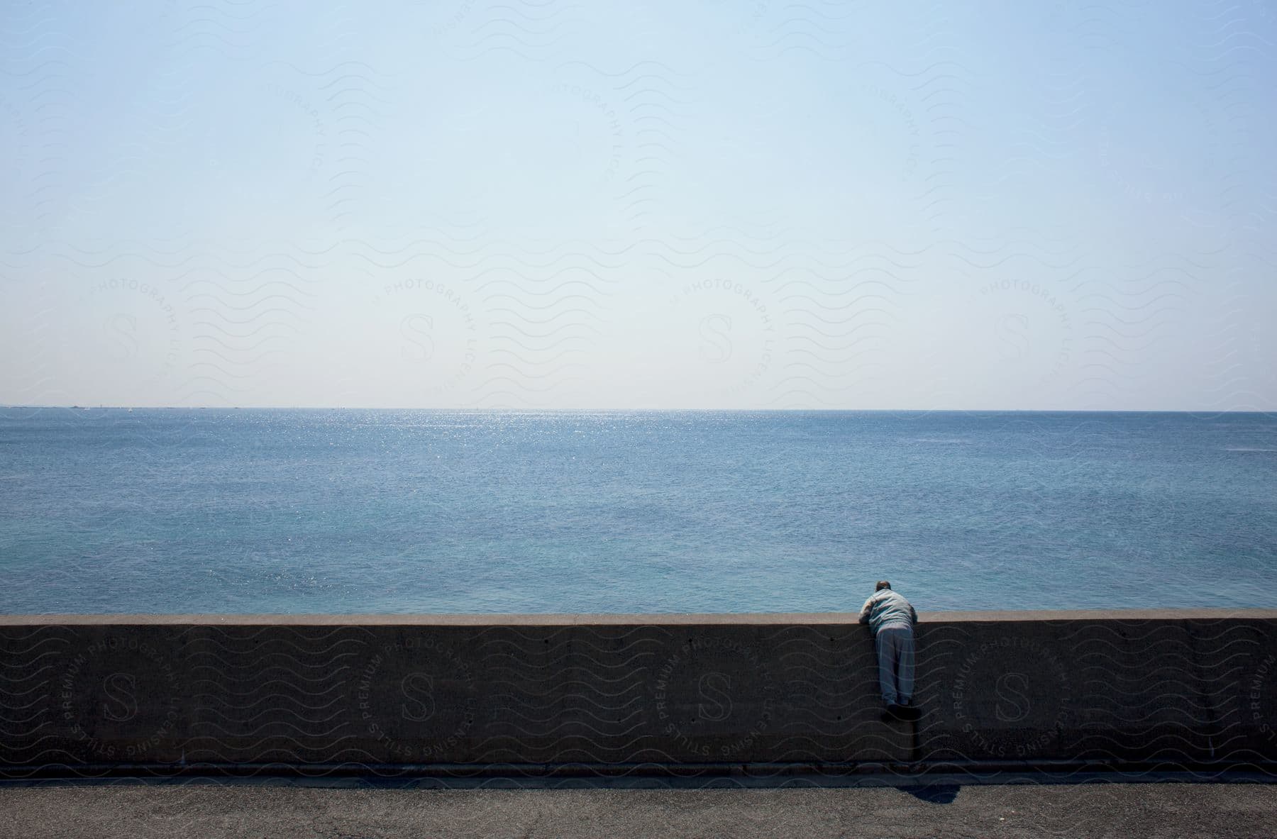 Man leaning over cement breaker wall facing open blue ocean under blue sky