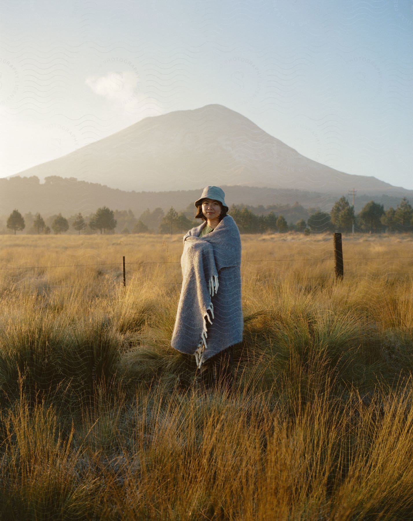 Woman in hat and blanket stands in grassy field with mountain in distance on sunny day