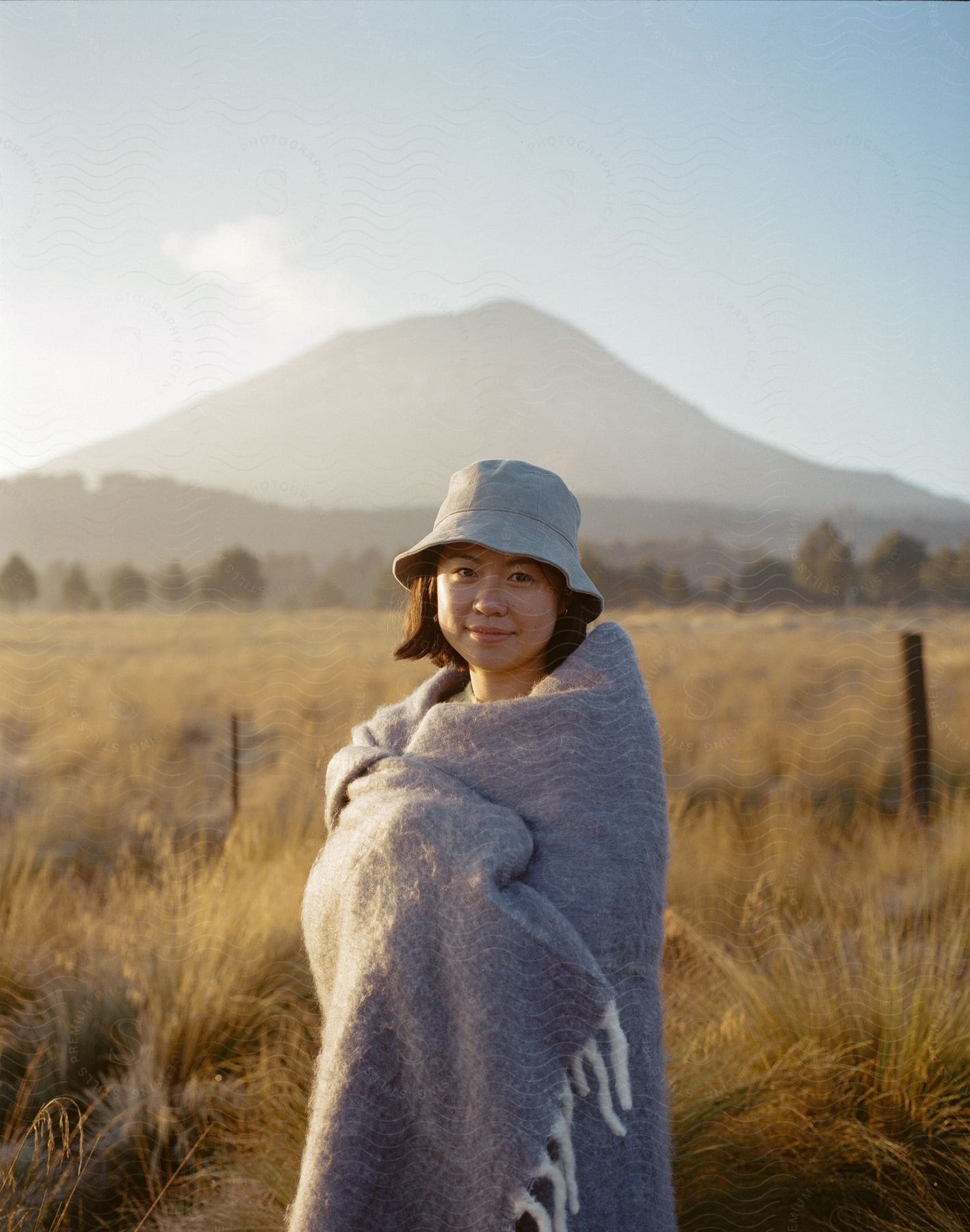 A young woman in a gray blanket stands in a grassy field with a mountain behind her