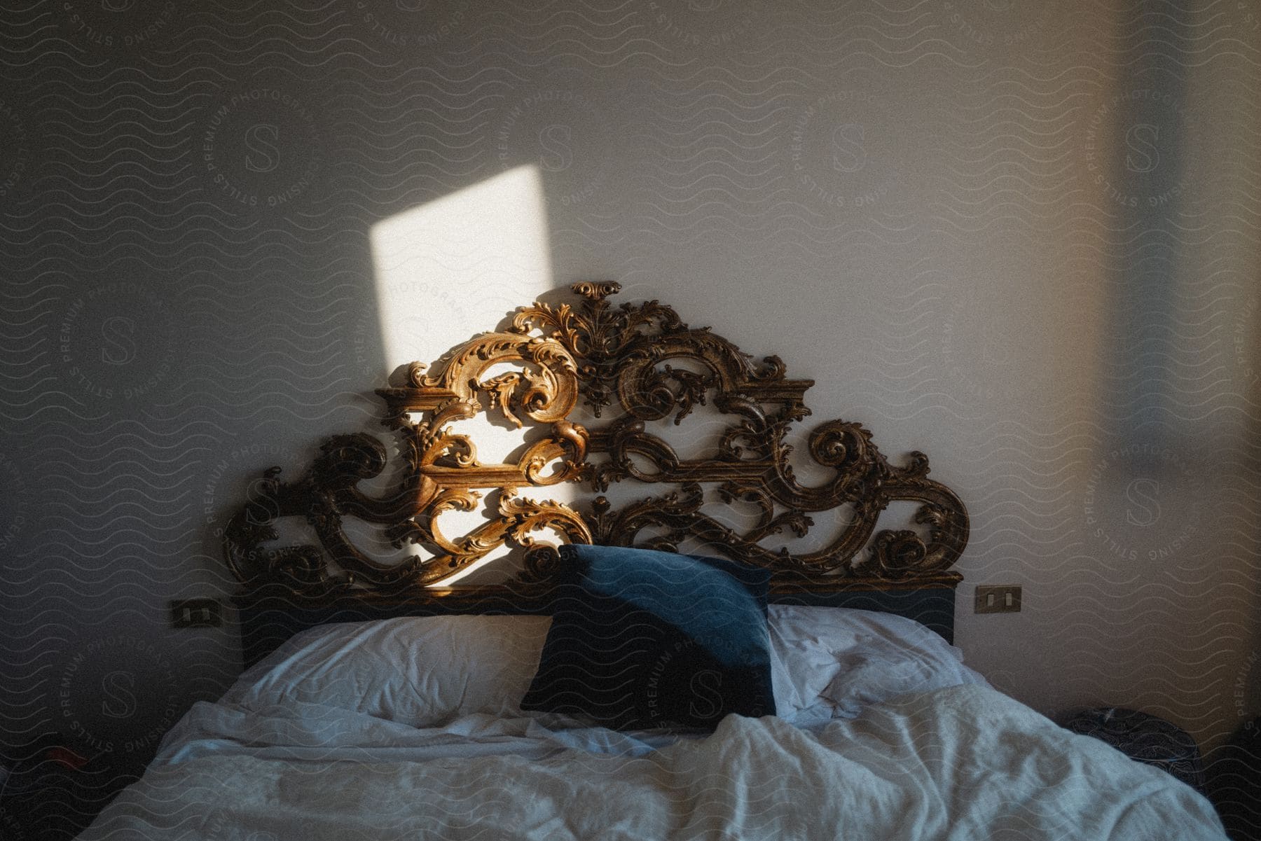 Blue pillow on bed with ornate headboard and sunlit rectangle on wall through window