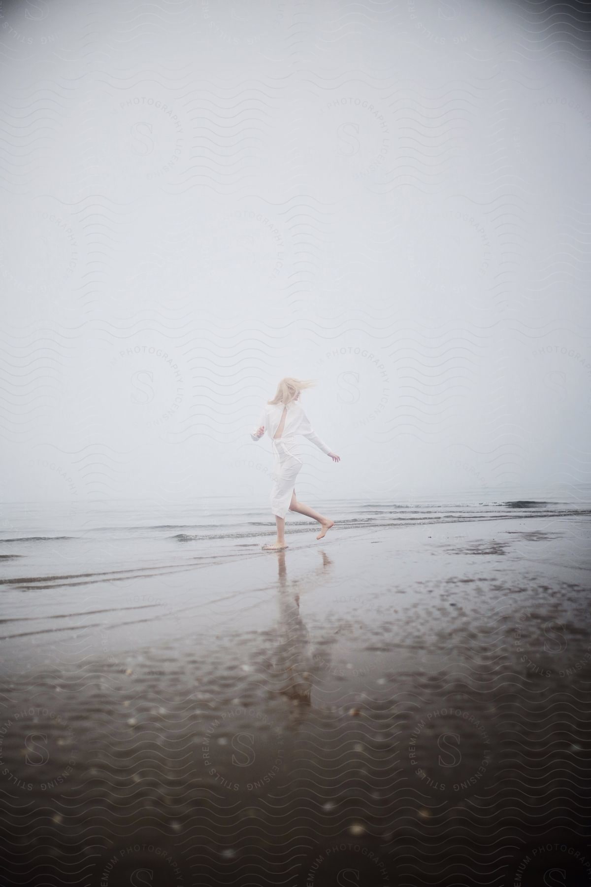 Blonde woman in white dress running on shoreline under hazy sky at beach