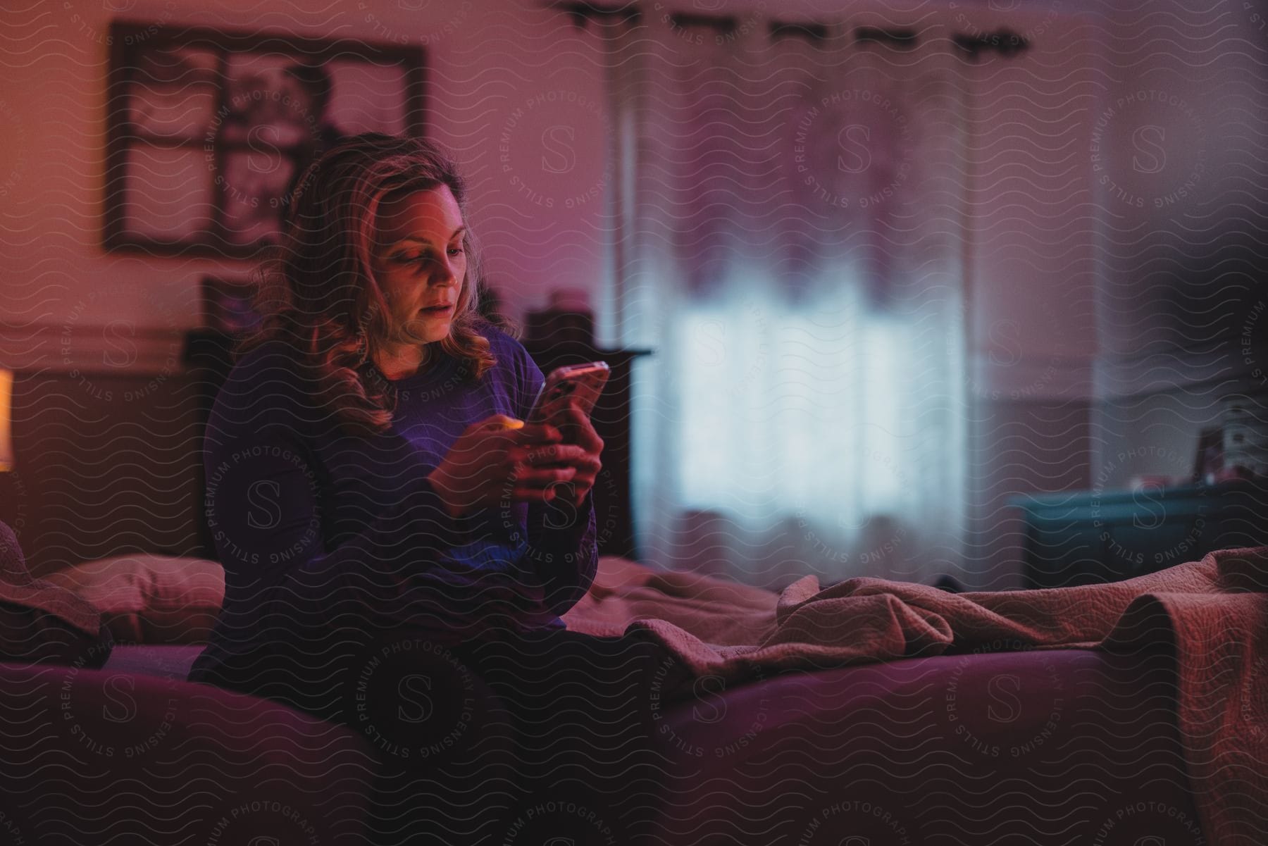 Blonde woman in sweatshirt and black pants sits on bed using smartphone in bedroom