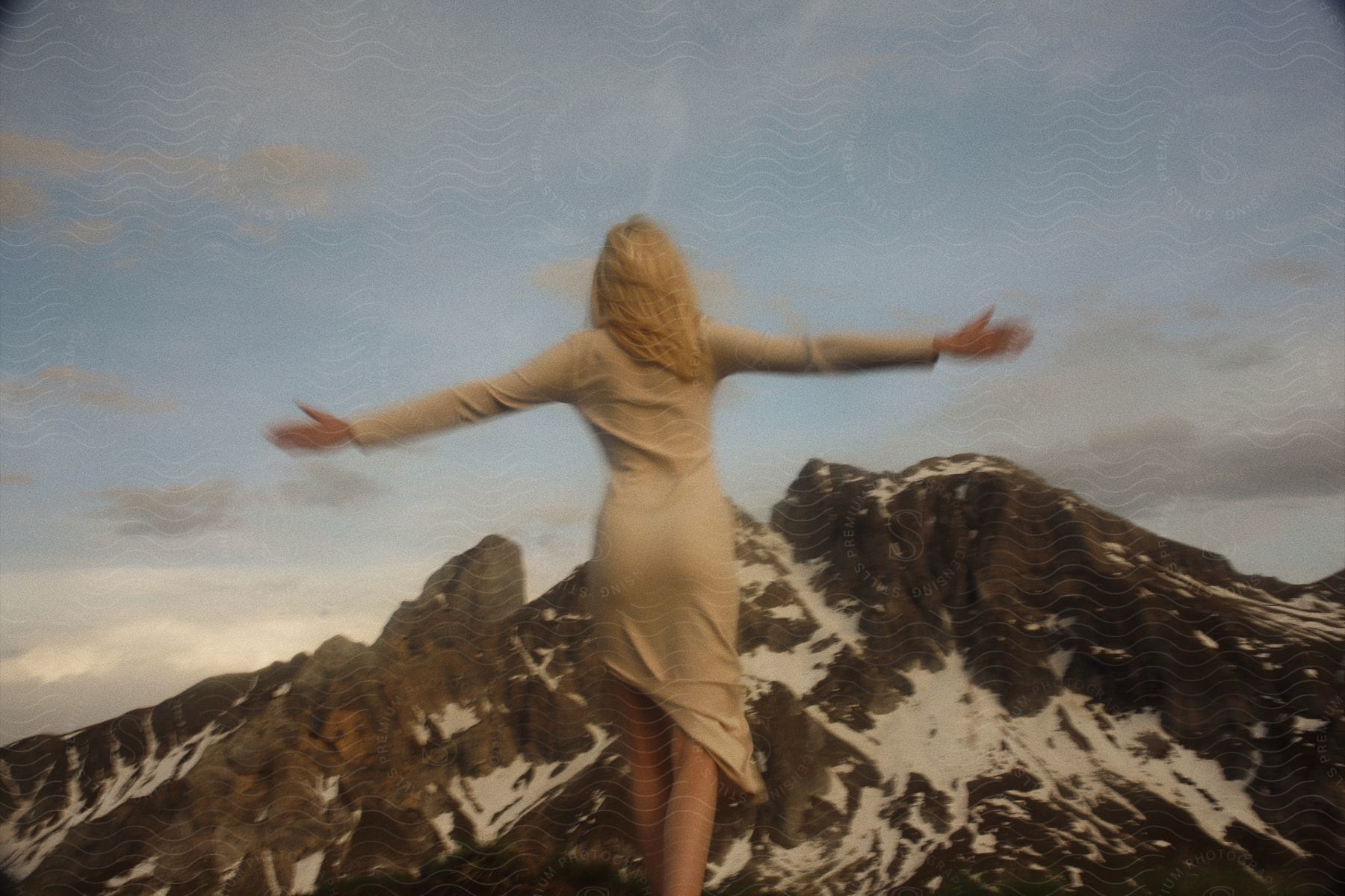 Blonde model in beige dress with outstretched arms in front of snowy mountain peak under dusky sky