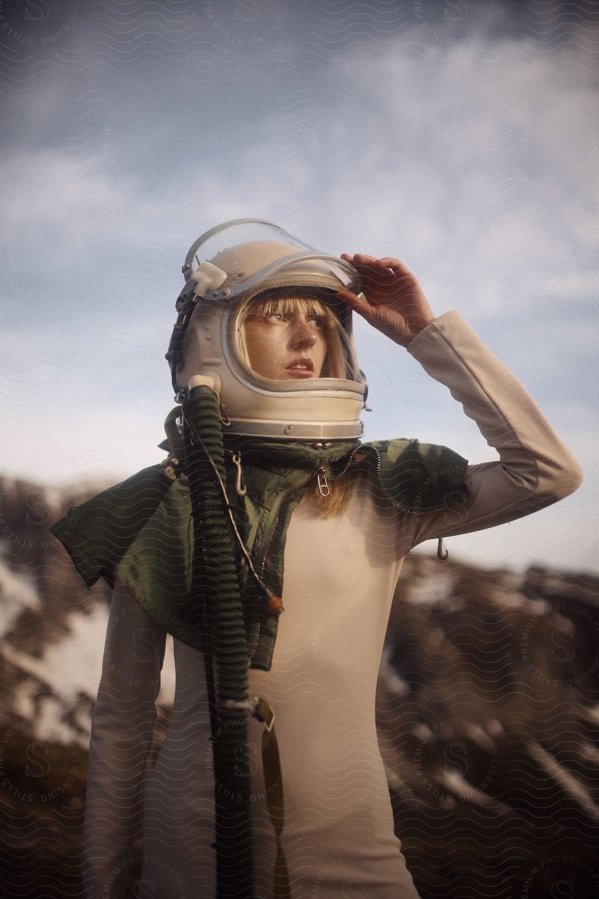 Young woman wearing a pilot helmet stands in front of the mountains looking at the sky at sunset