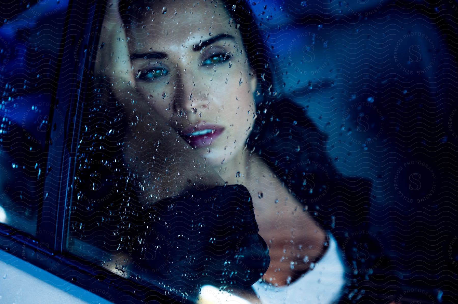 Darkhaired woman posing inside a car seen through the window covered in water drops