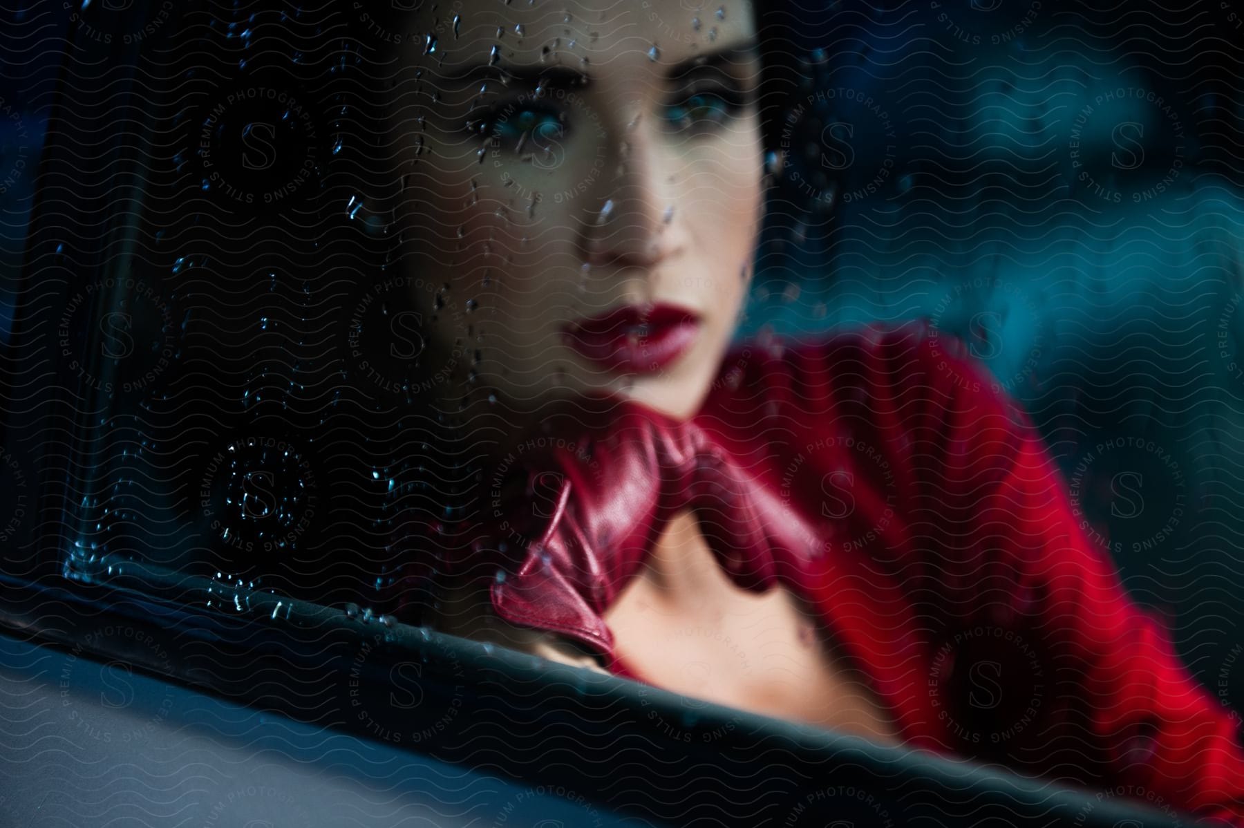 A woman in a red dress rests her chin on her hand while sitting in a black car on a rainy day
