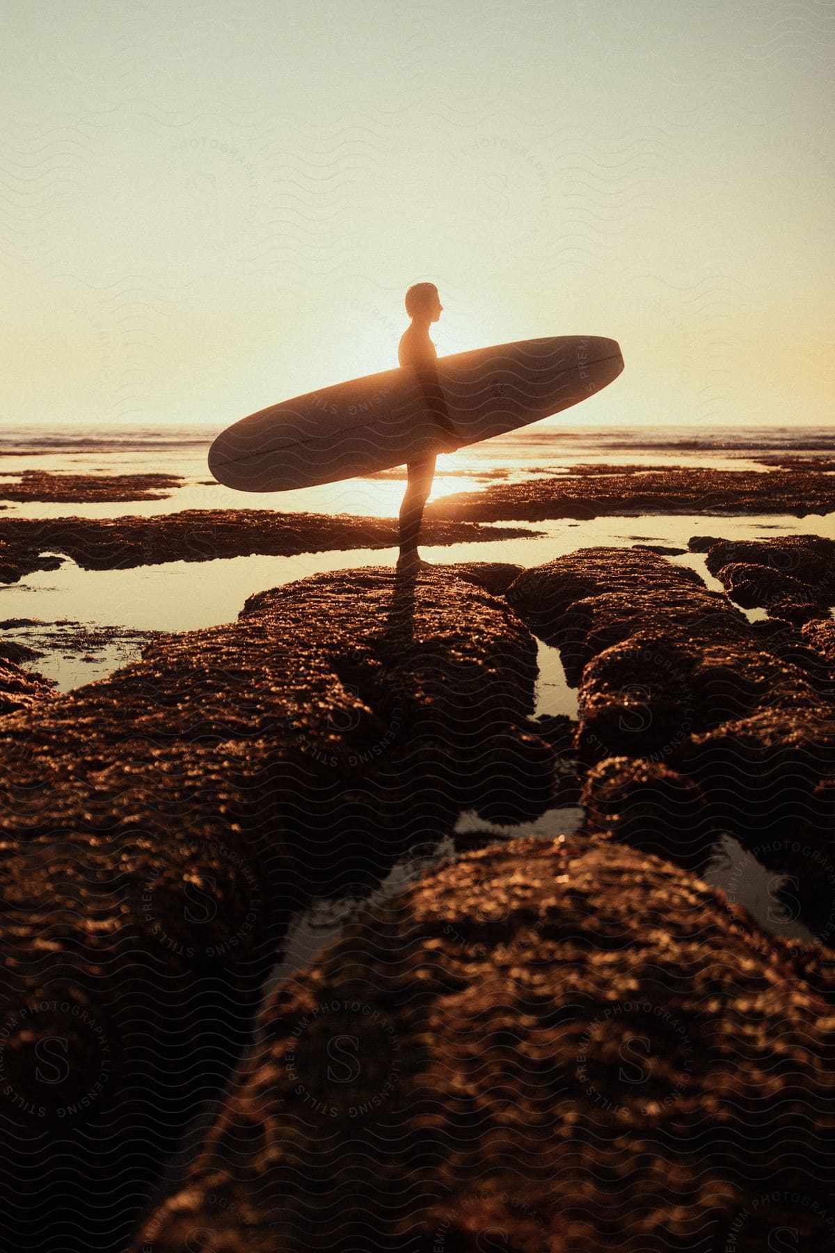 A man holding a surfboard is standing on a rocky beach at sunset.