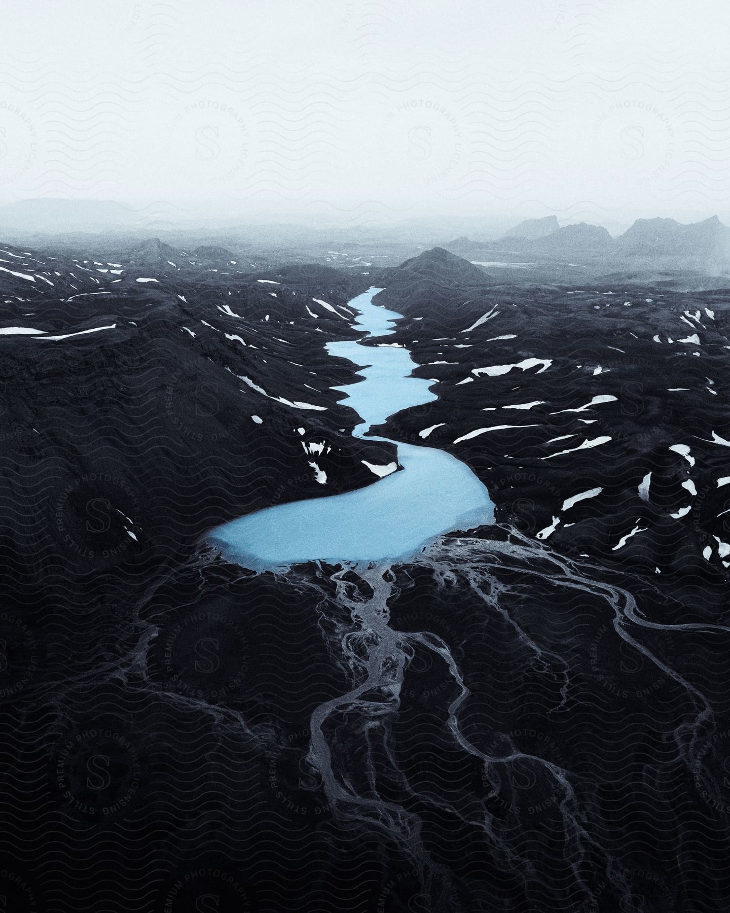 Bright blue river flows through black snow dappled terrain on a foggy day.