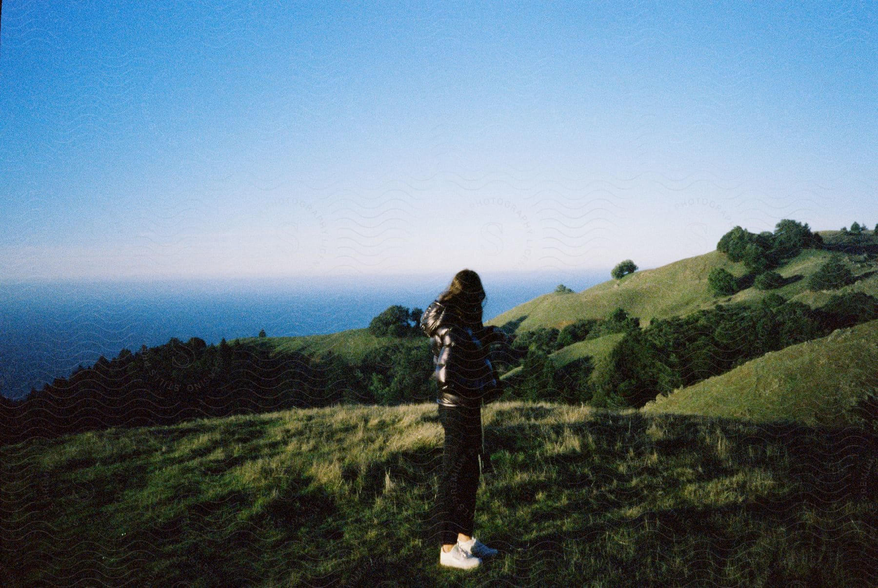 A woman wearing a black coat black pants and white shoes stands on a grassy hill near the ocean