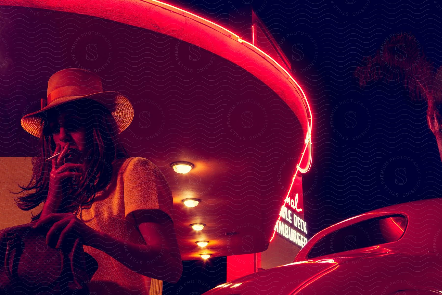 A woman in a straw hat smokes next to a car at a hamburger restaurant lit by neon