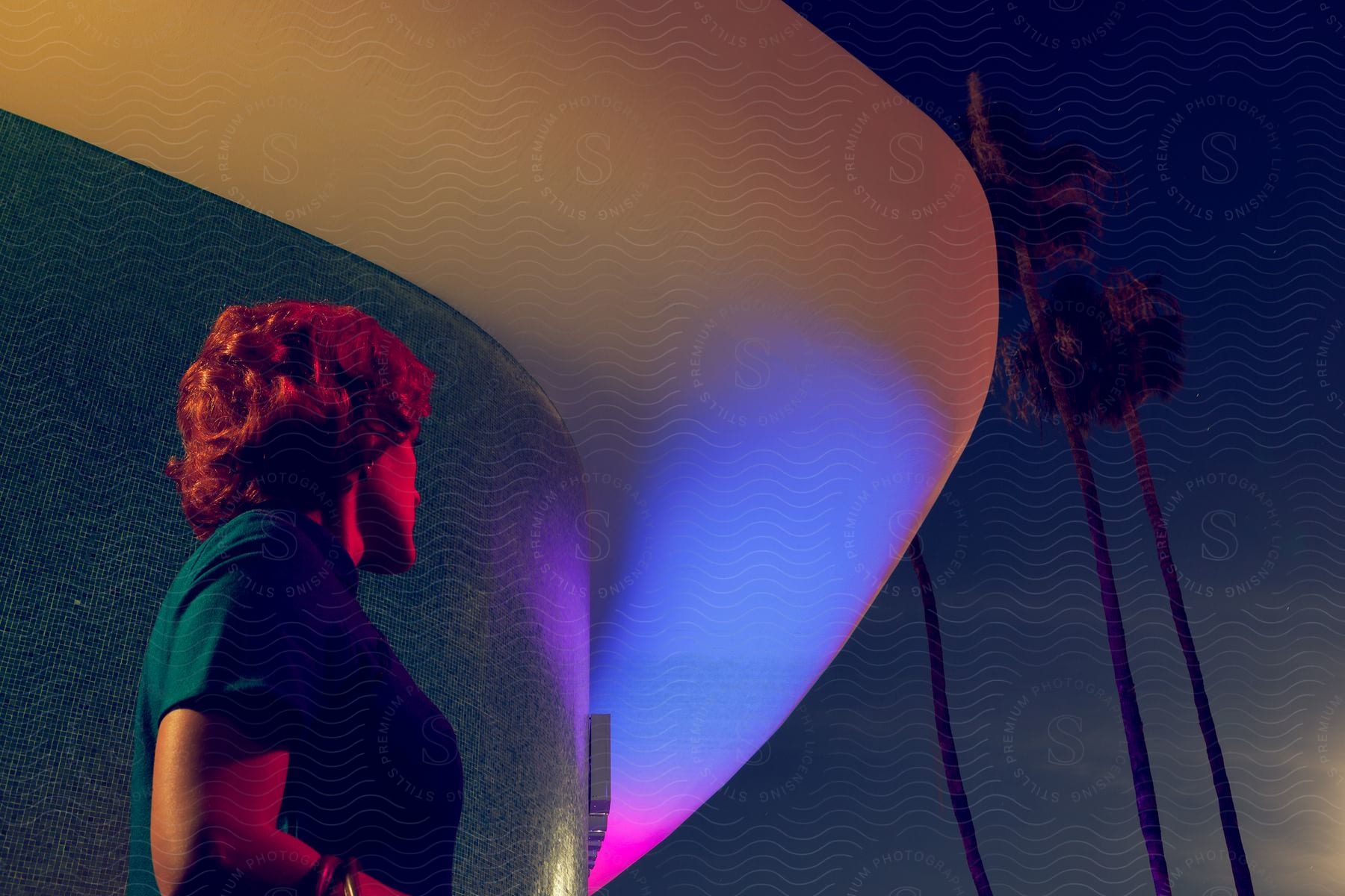 A woman with 70s style hair stands in front of a retro building at night with tall palm trees extending above a curved overhang