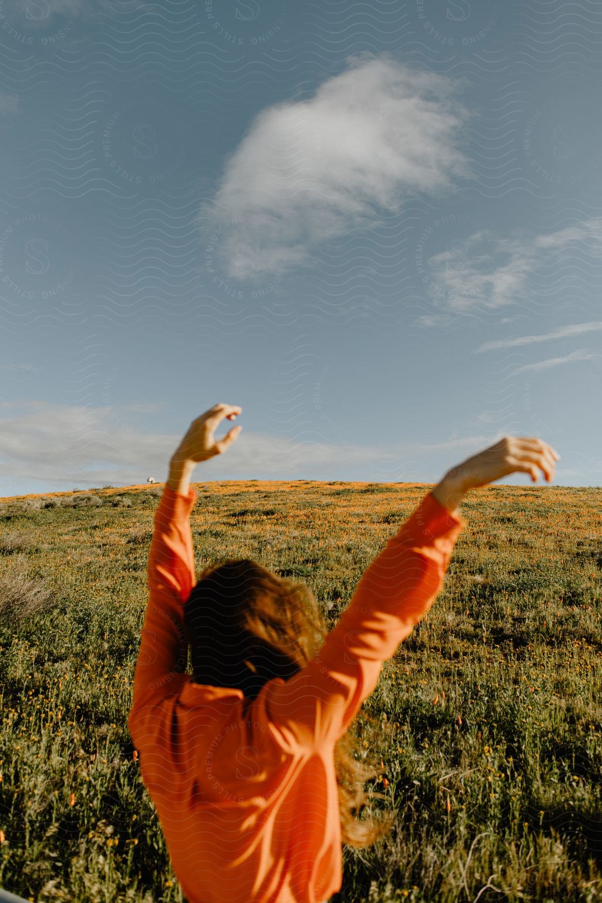 A woman stands in a green field raising her arms towards the sky as clouds float above