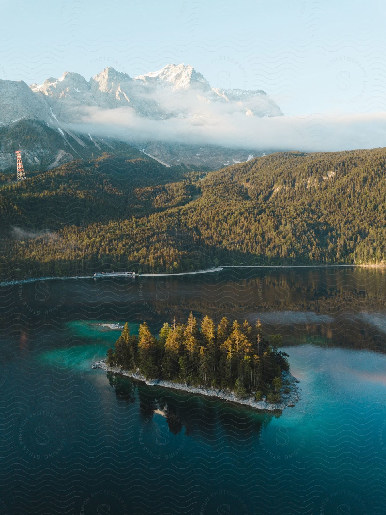 A serene lake in springtime with blossoming trees and a small island framed by a cell tower and a snowcapped mountain in the distance