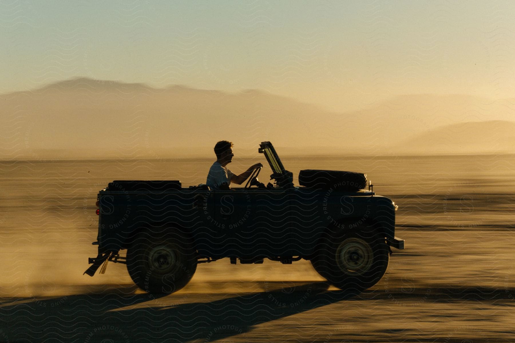 Man driving a jeep in the desert at sunset kicking up dust with the wheels leaving a dust cloud behind