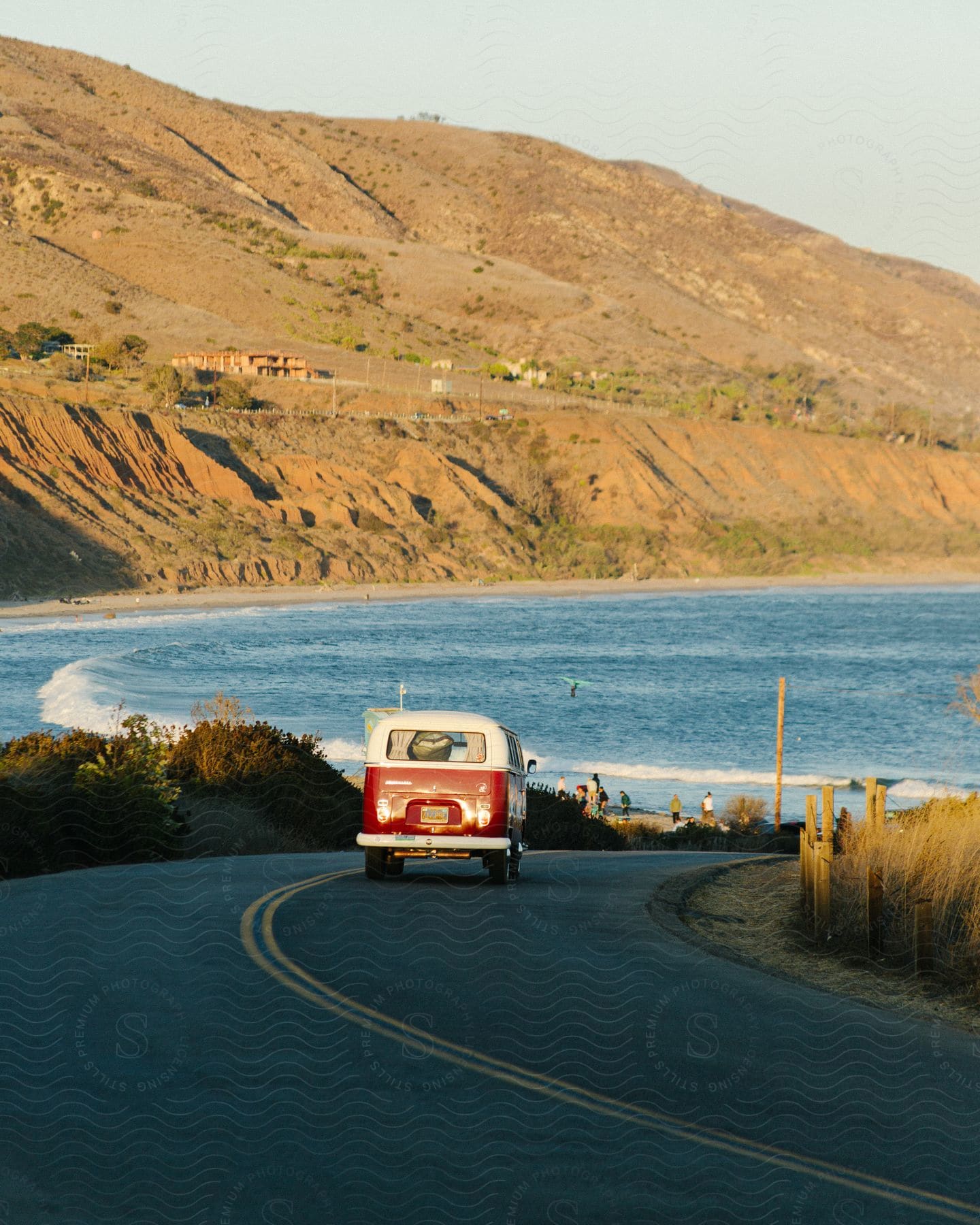 An Old Red Van Driving Down A Coastal Road With Waves Crashing Ashore And Mountains In The Distance