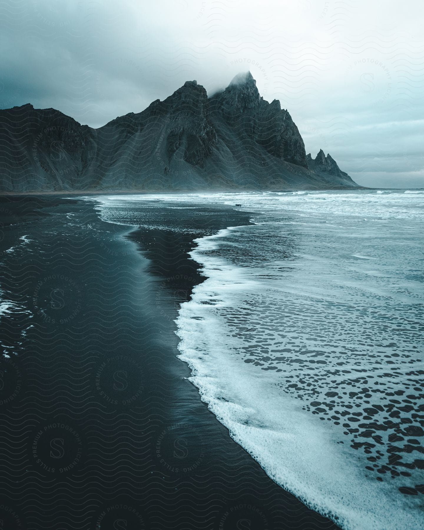 Ocean waves crashing onto a dark sandy beach with distant pointy mountains and fog settled over the ridges