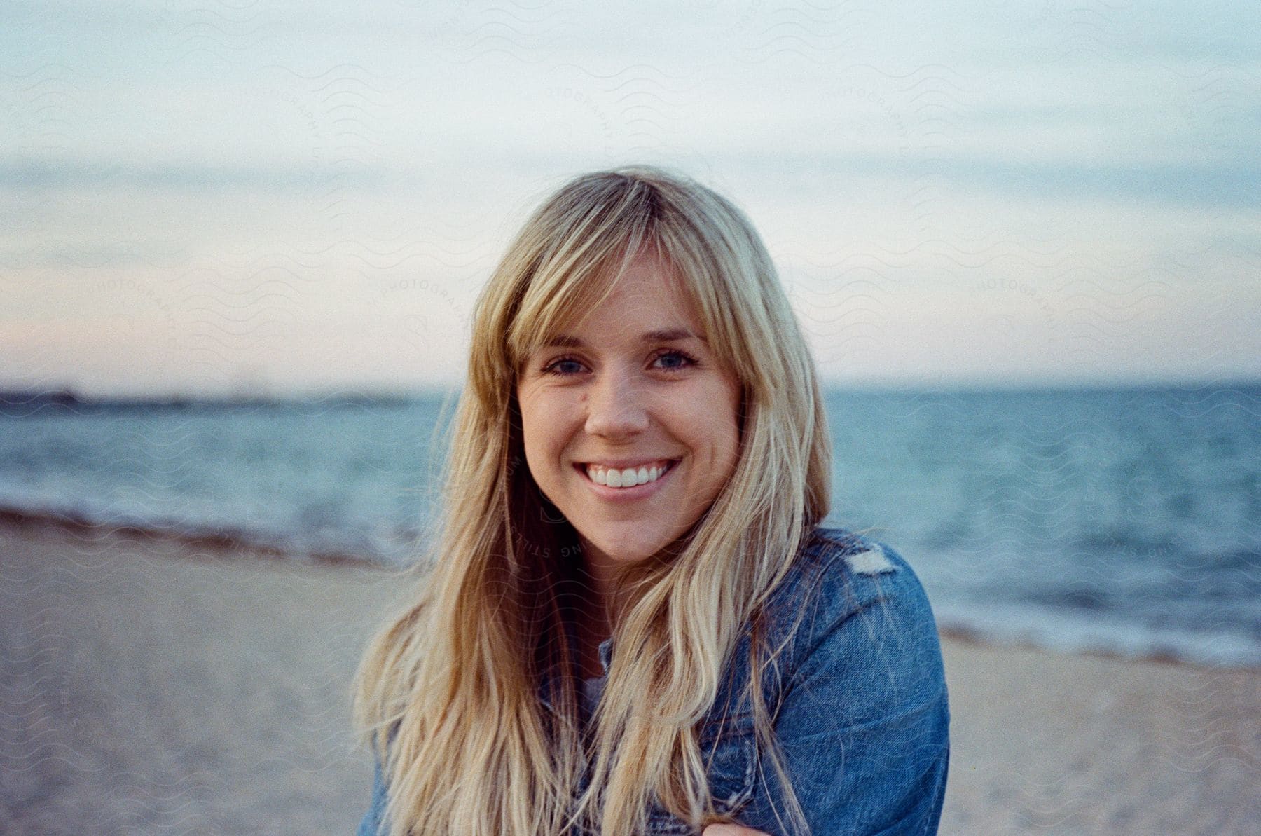 Blonde woman wearing a denim jacket stands on sandy beach next to the ocean