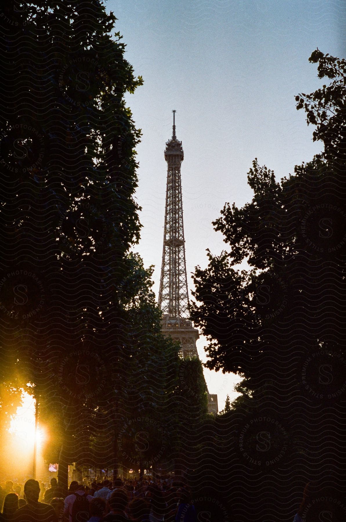 Sunlight breaks through the trees during sunset in a crowded park with the eiffel tower in the background
