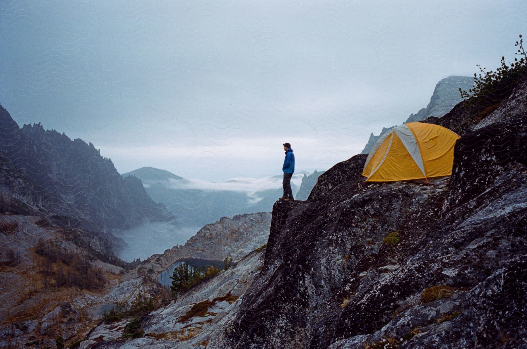 A man stands on the edge of a mountain cliff in front of his tent