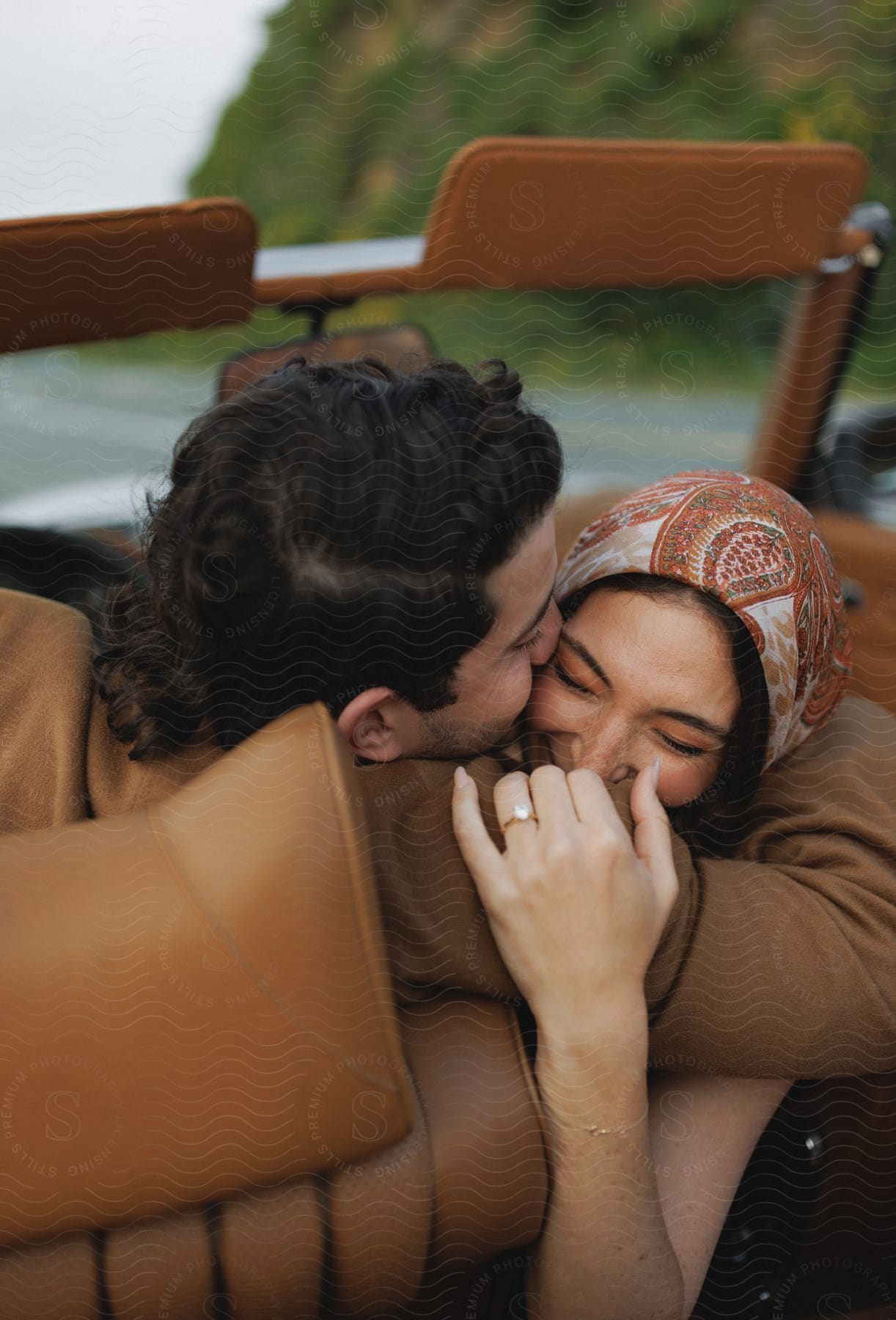 A man and a woman in a convertible embrace each other smiling and sharing a kiss on the womans forehead