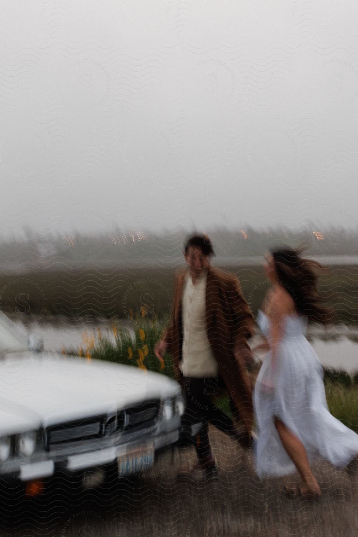 Man in brown jacket and white shirt holds womans hand in wedding dress walking to white car with city skyline in background in field