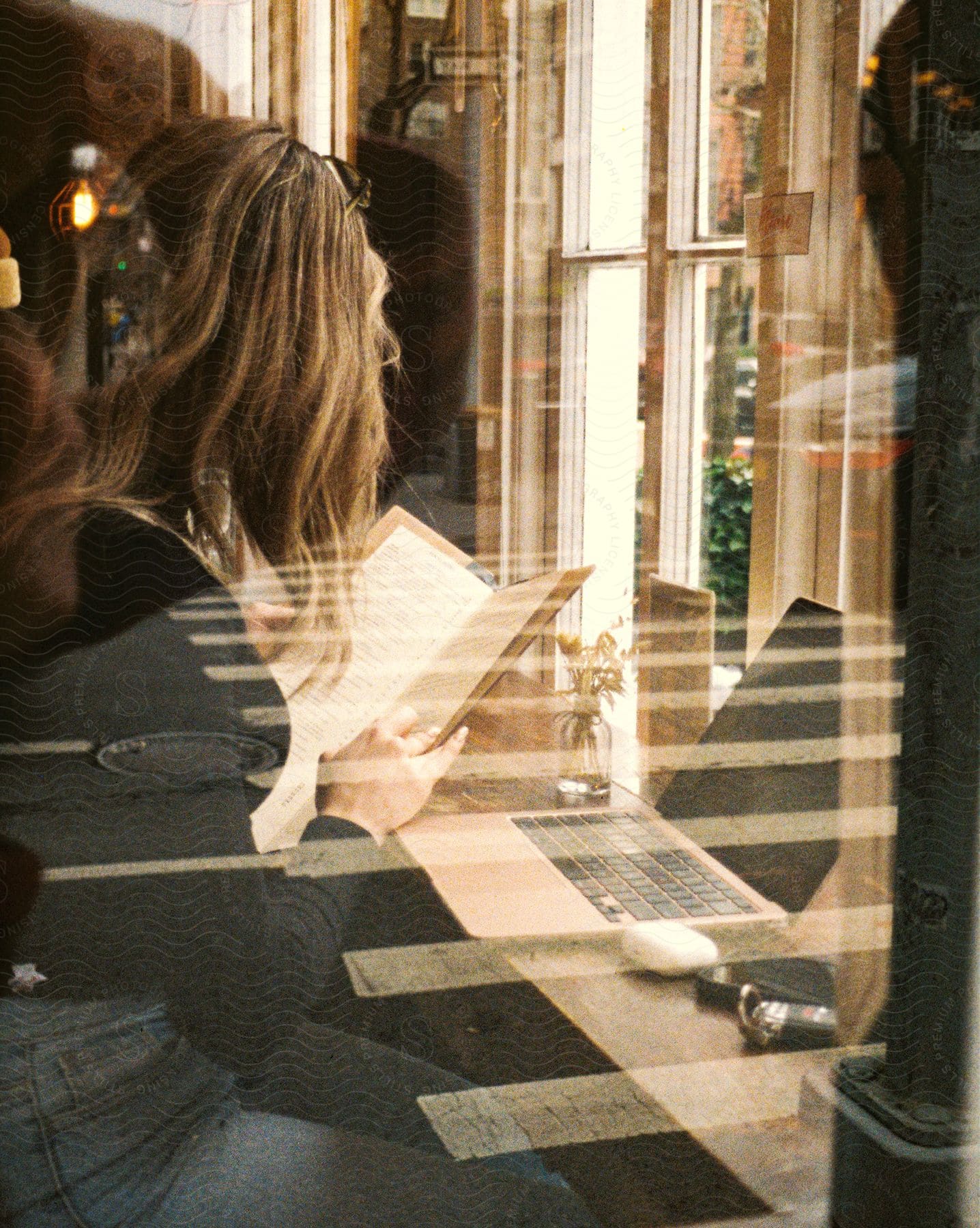 Stock photo of a woman is sitting at a table in front of windows with a laptop computer as she looks at a book