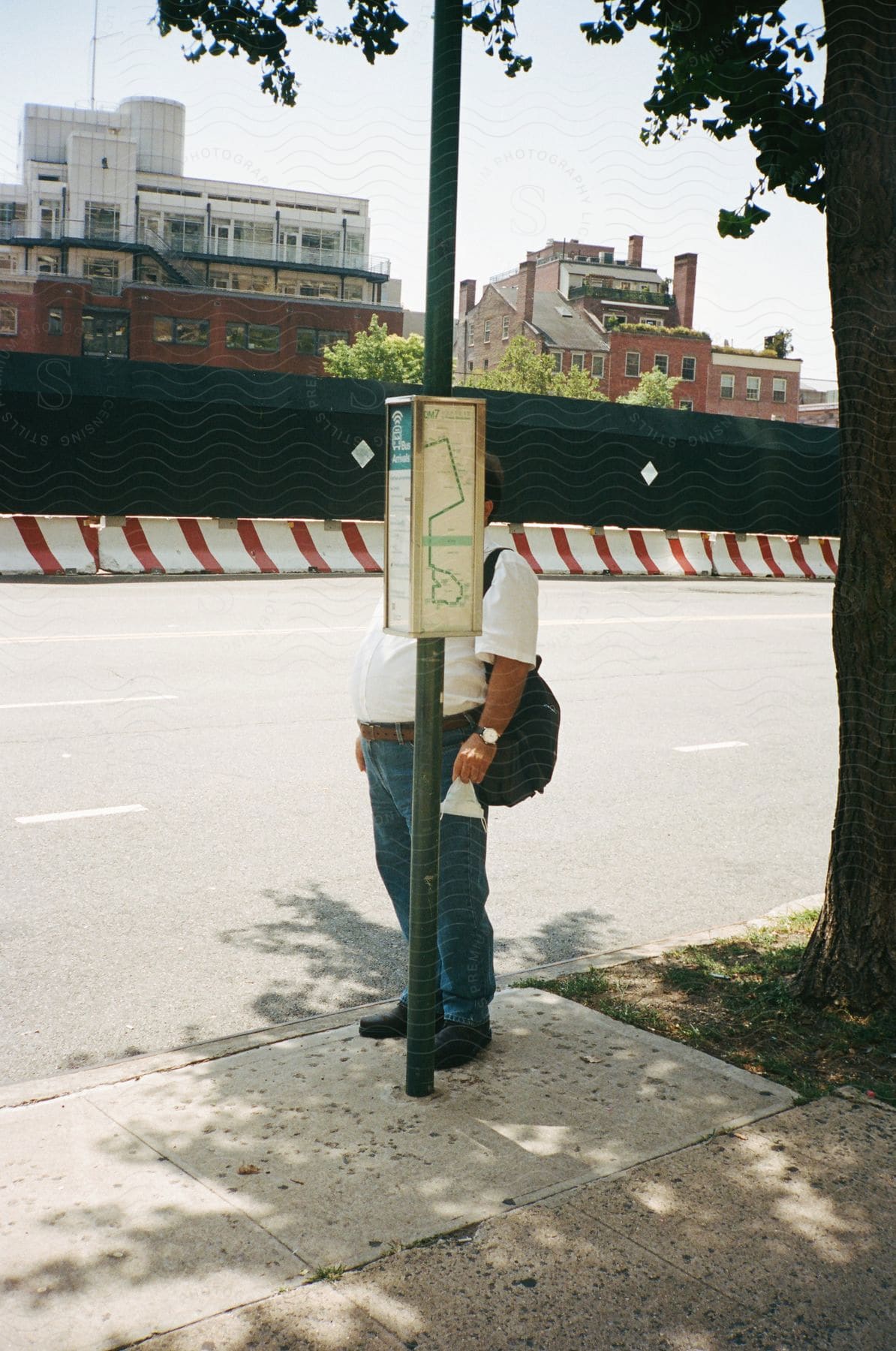 A man carrying a backpack stands behind a street pole, hiding his face at mid-day