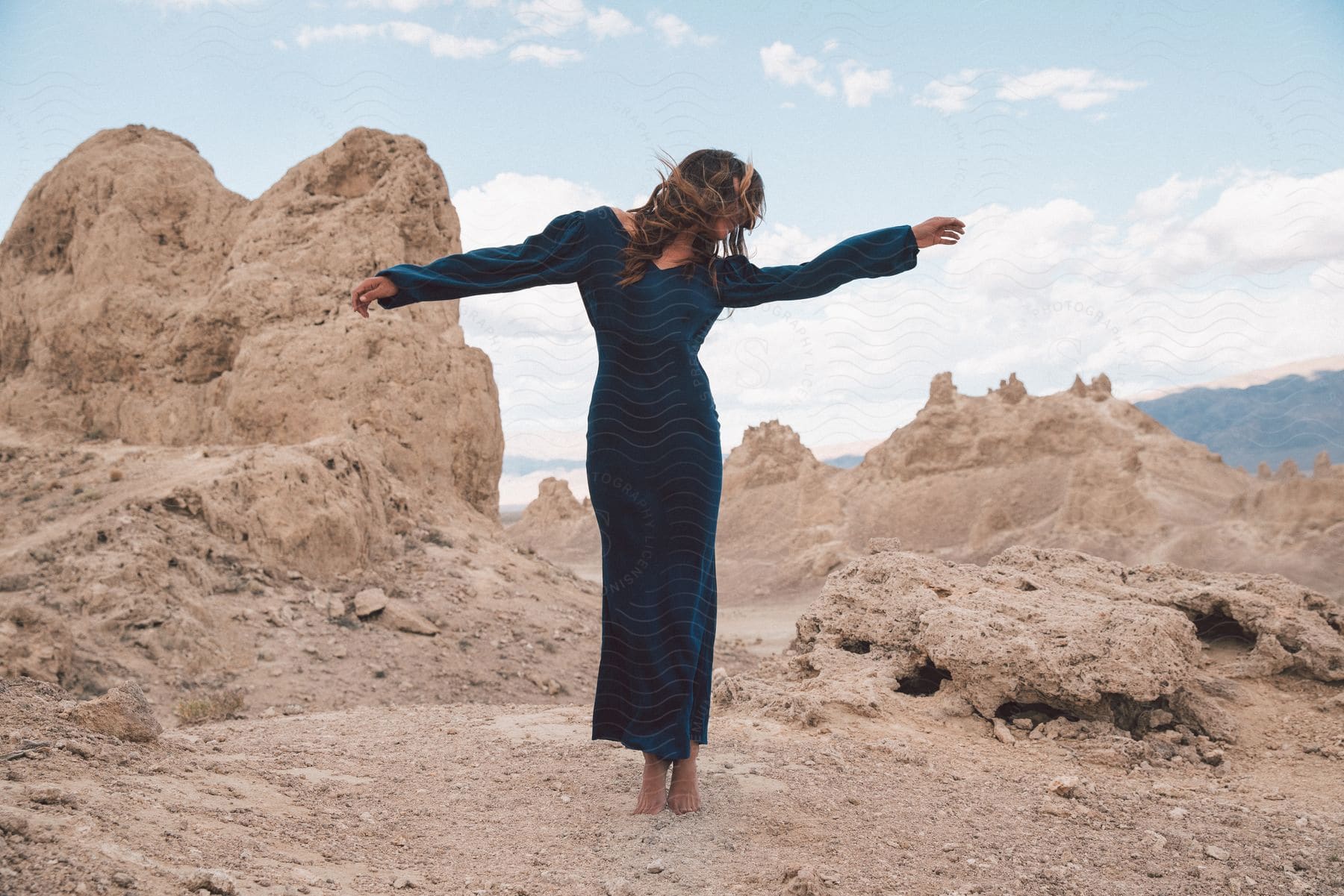 A woman in a black dress dances among rock formations on a desert mountain with distant mountains