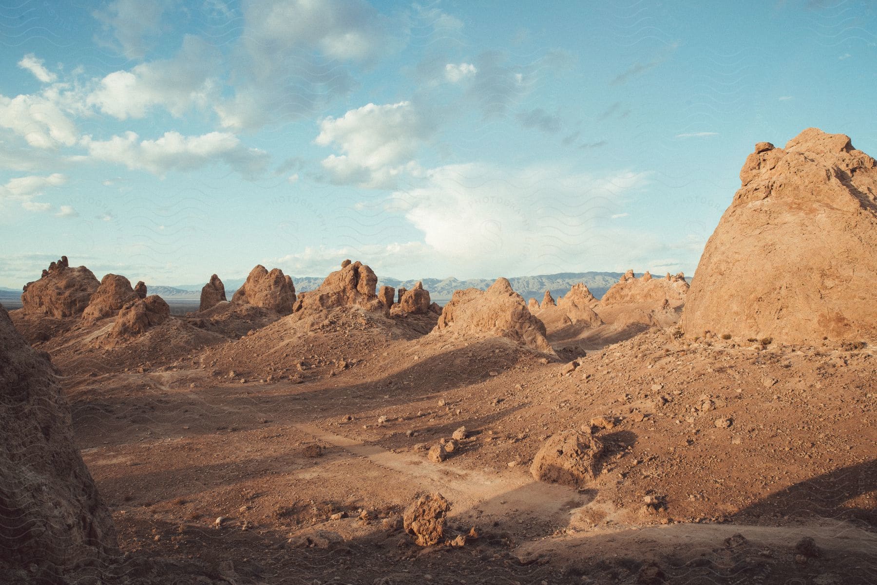 A trail winds through desert badlands with sunlit buttes and rock formations under a cloudy sky