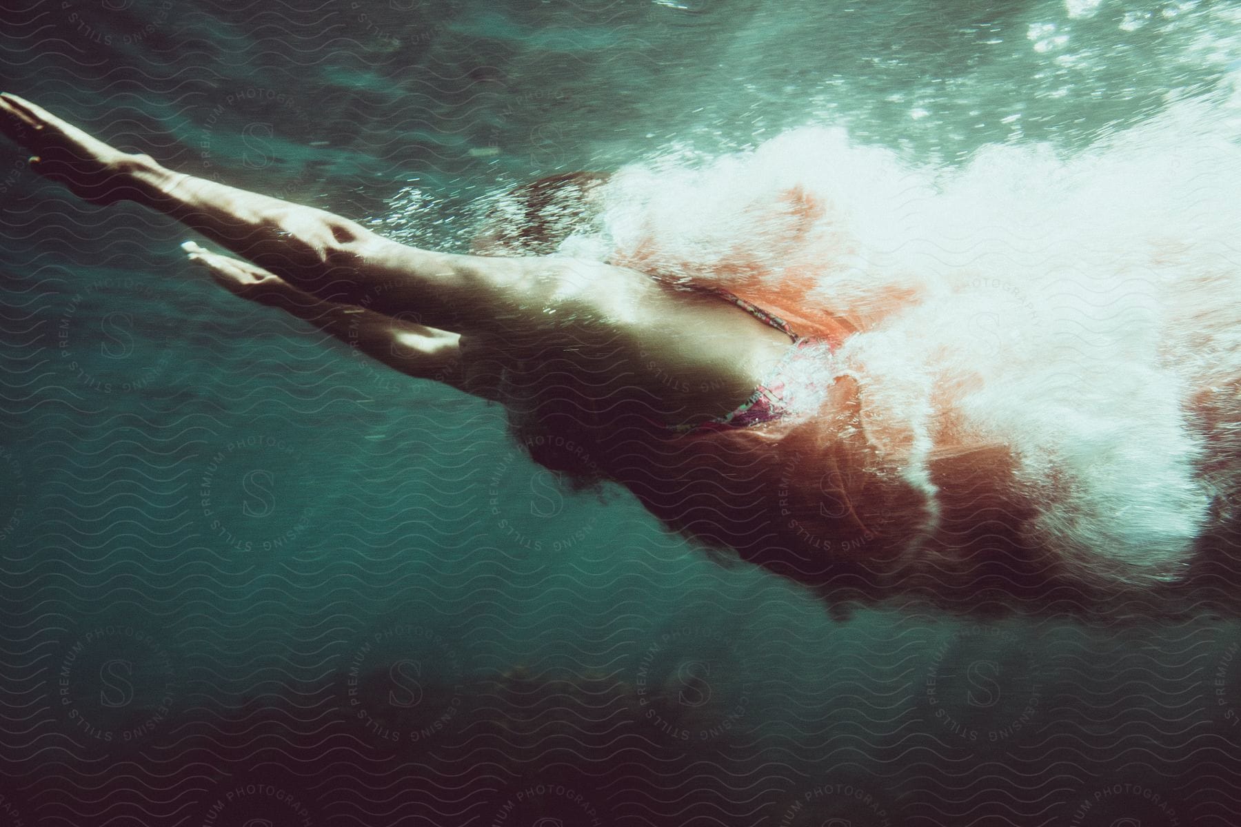 A woman swims underwater in the dark ocean with sunlight illuminating her body