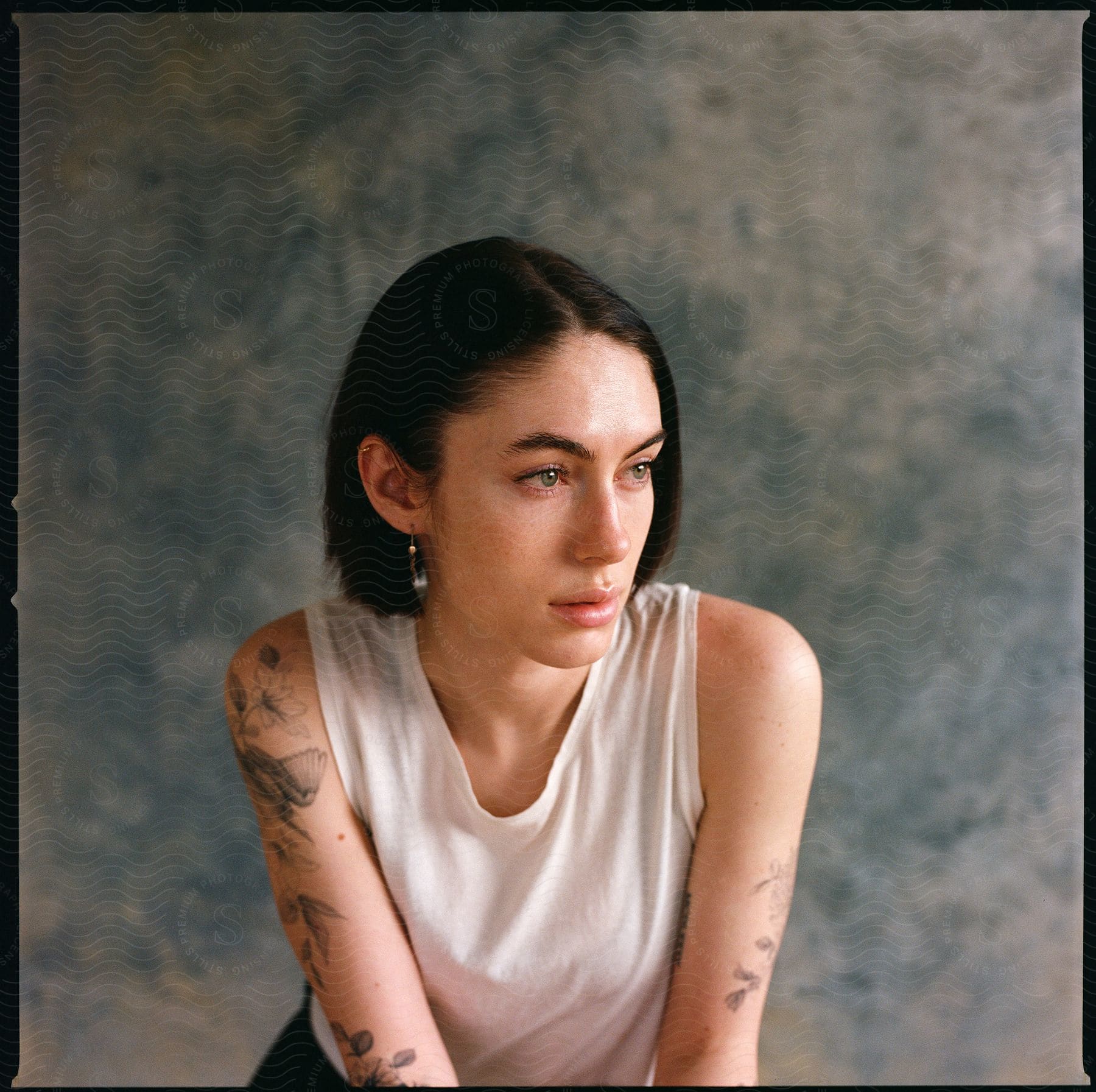 Short haired woman with tattoos and white tank top looking off to the side indoors