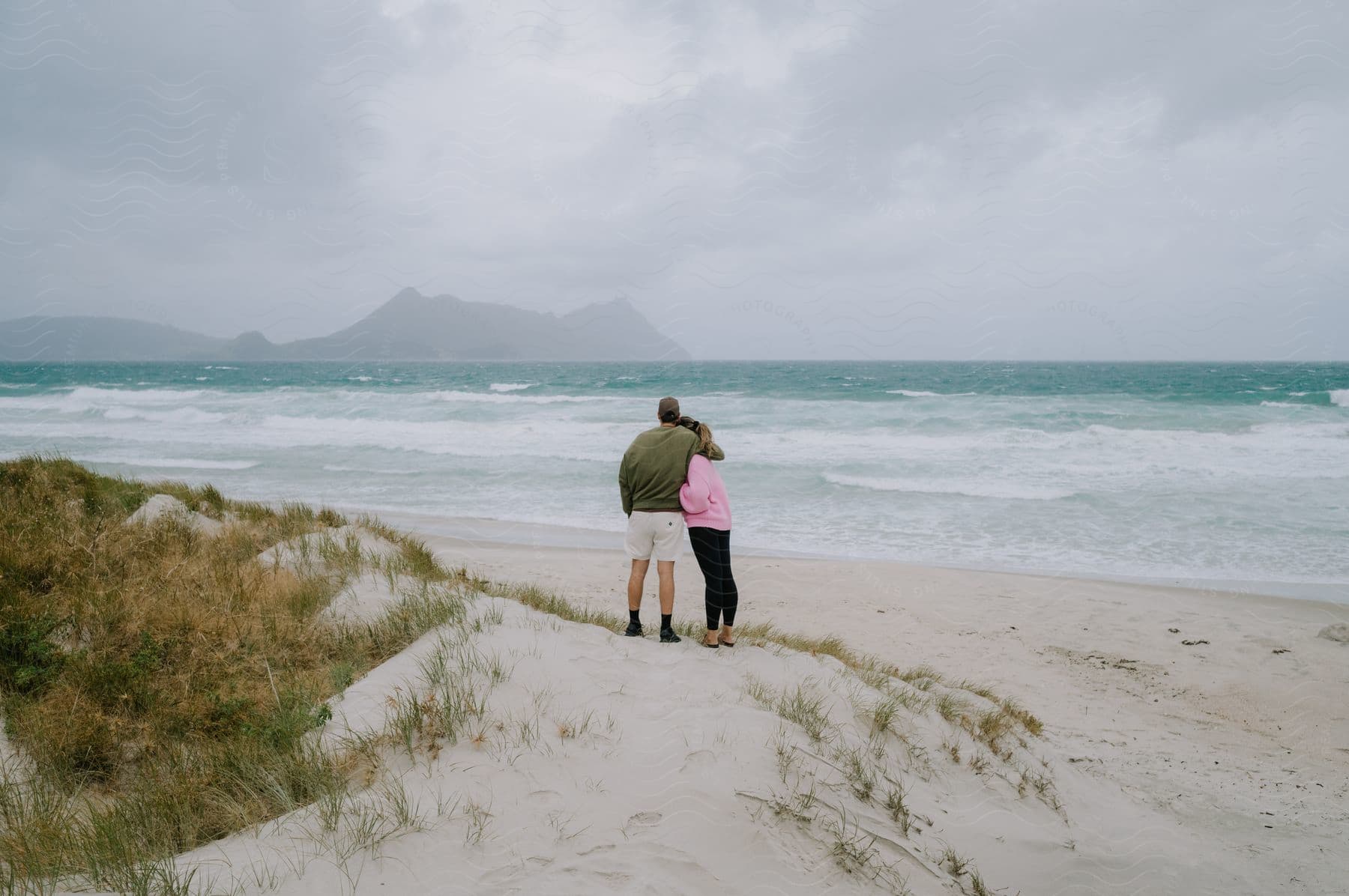 Couple hugging on sandy beach admiring turbulent waves in ocean