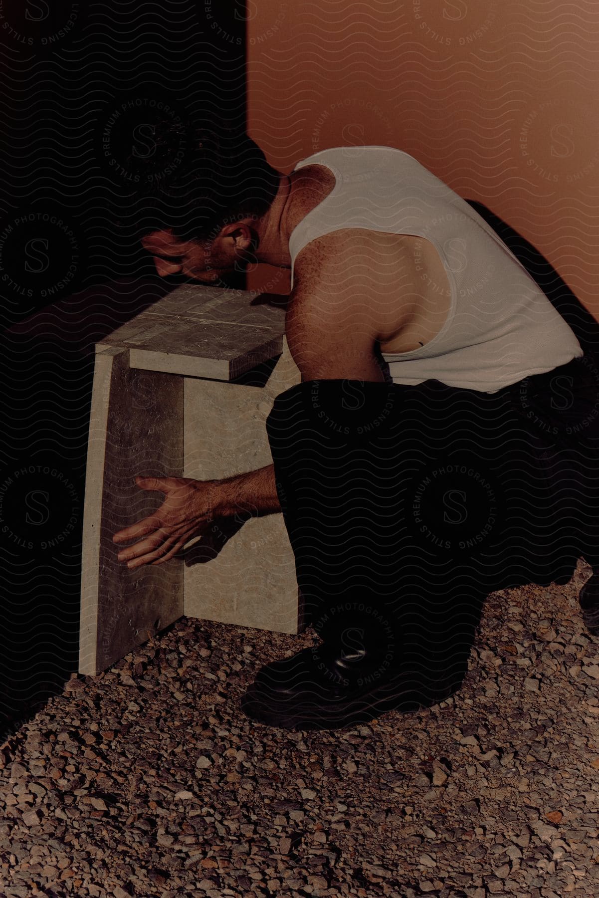 A determined adult man sitting on a gravel road wearing pants and an undershirt