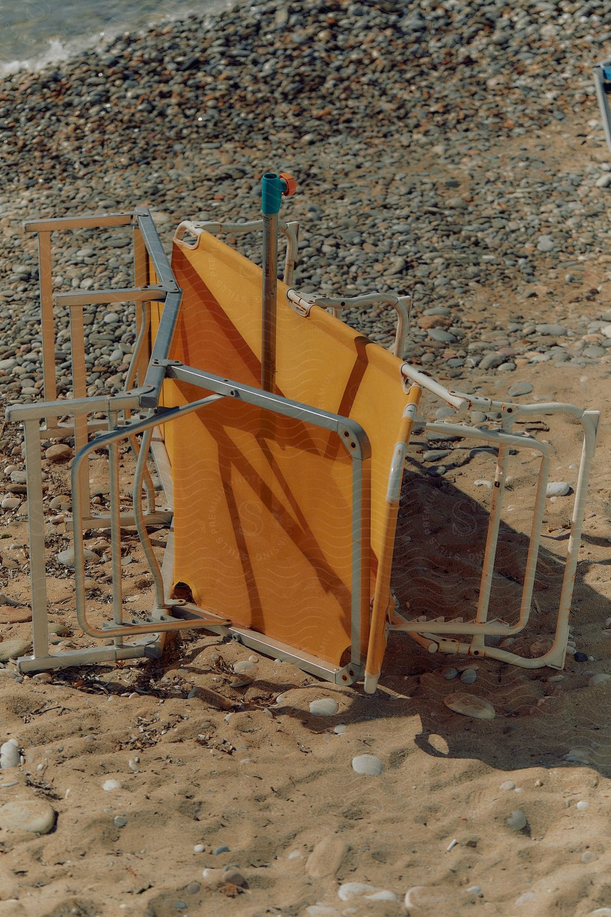 A chaise lounge beach chair lays on its side in between an umbrella pole on a rocky beach