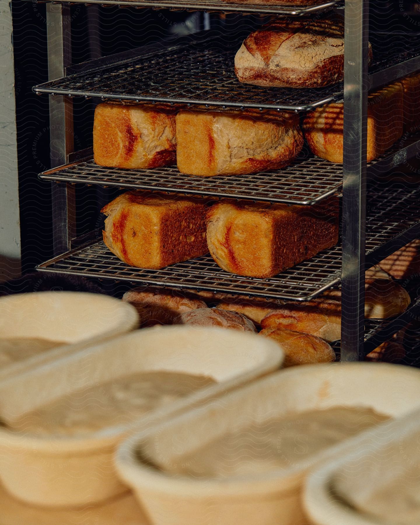 A metal cooling rack filled with fresh bread loafs in a bakery with other pans of dough lined up for baking