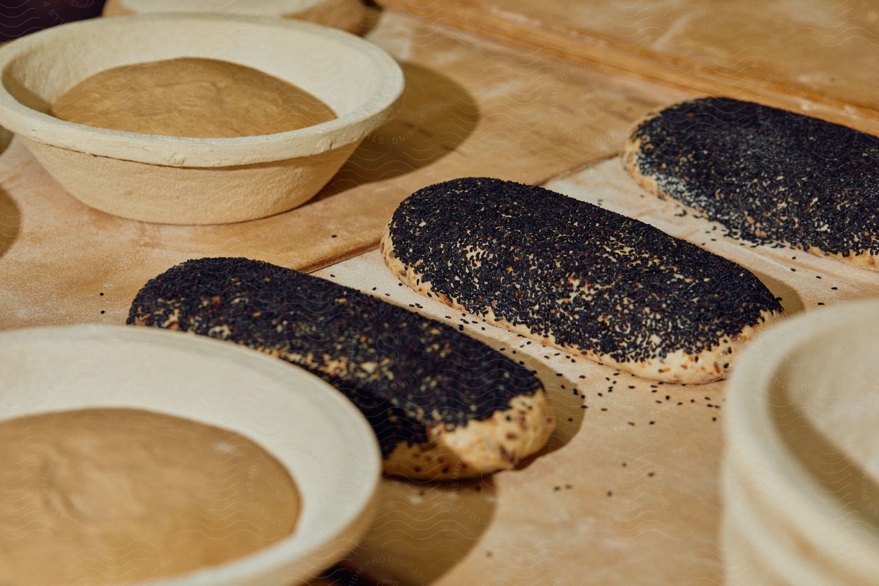 Bread covered in black sesame laid over a wooden table with other bread being prepared around in an industrial kitchen