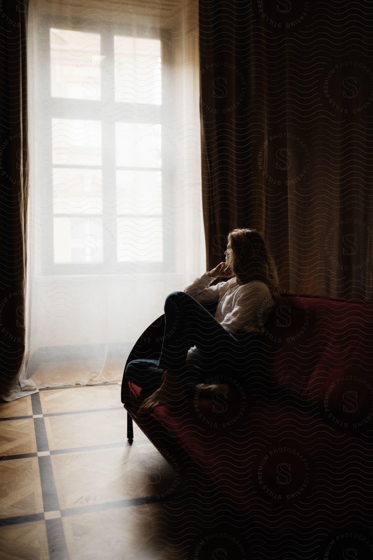 A woman sitting in a large chair stares out a window in a dark room