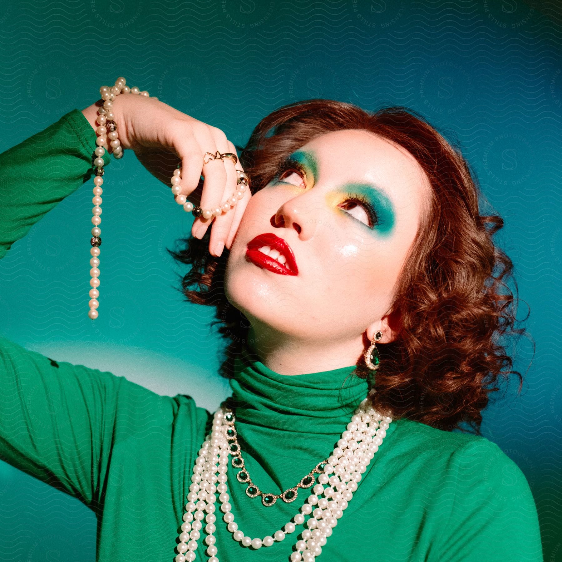 A redhaired female model wearing a green turtleneck and pearl jewelry poses with her head tilted up and to the right her right elbow above her shoulder and hand touching her cheek