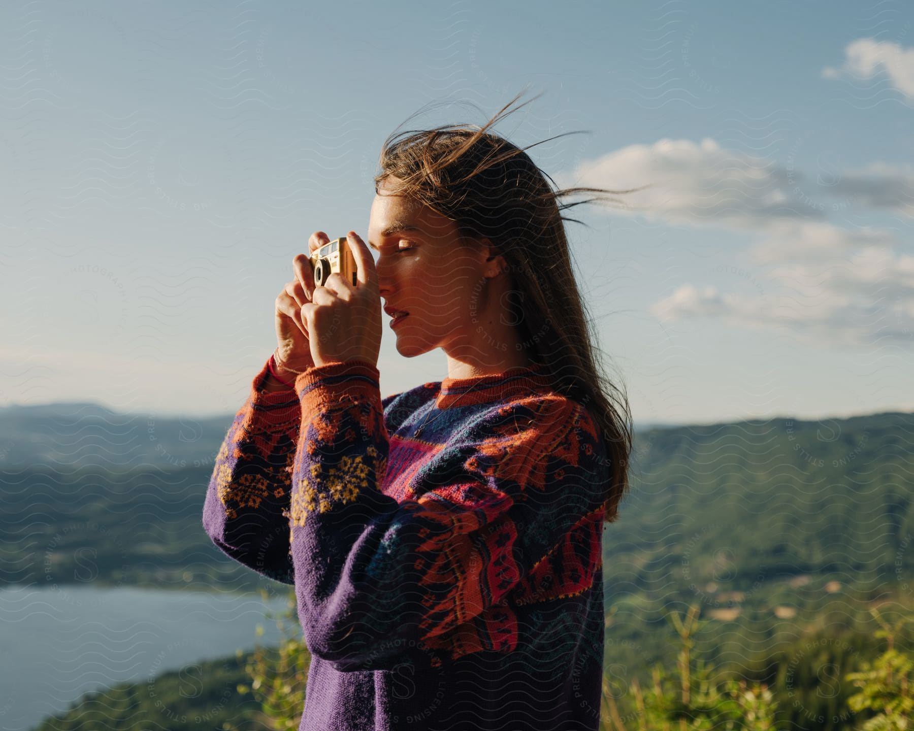 Stock photo of teenage girl in sweater holding camera with wind blowing her hair standing over mountain river valley